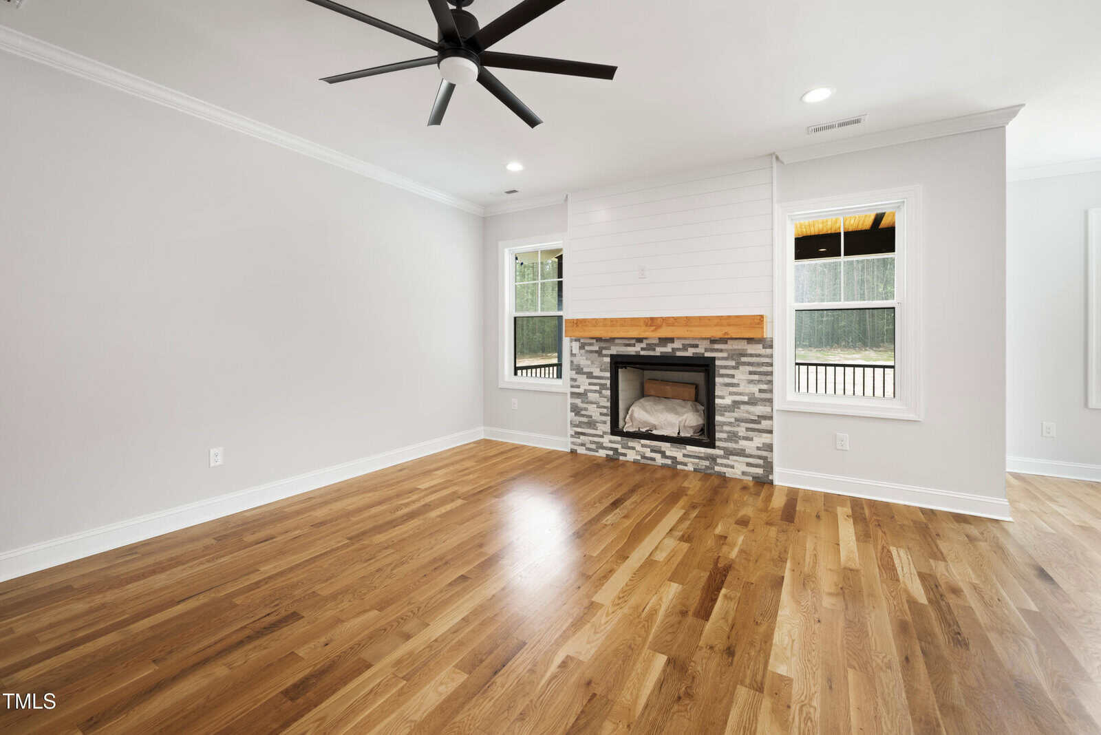 A bright, empty living room with light-colored walls, hardwood floors, two windows, a ceiling fan, and a fireplace featuring a stone surround and wooden mantel.