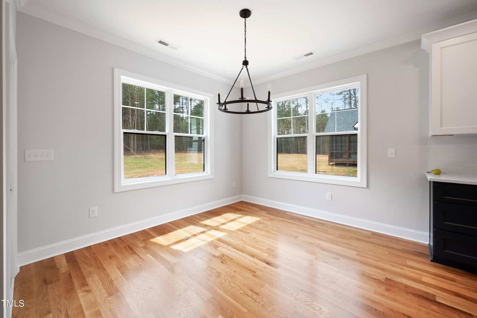 A bright, empty room with light gray walls, two large windows, hardwood floors, and a black chandelier hanging from the ceiling. Sunlight streams in, creating a warm atmosphere.