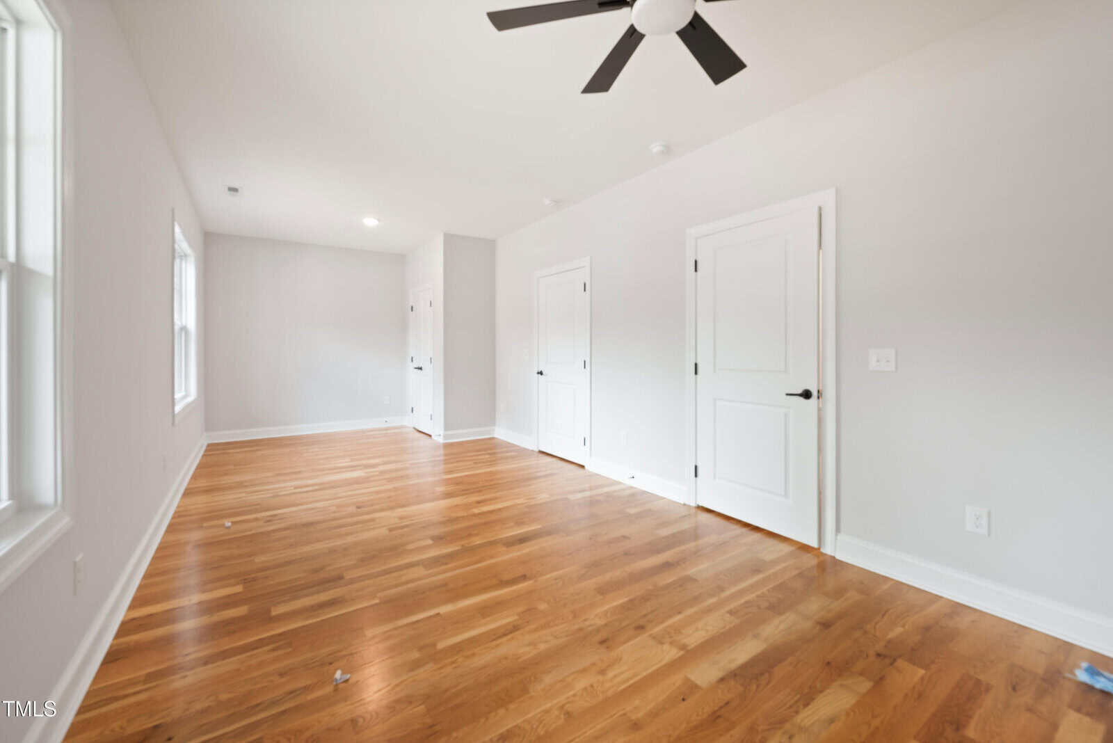 Bright, empty room with light-colored walls, hardwood floors, three white doors, ceiling fan, and large windows letting in natural light.