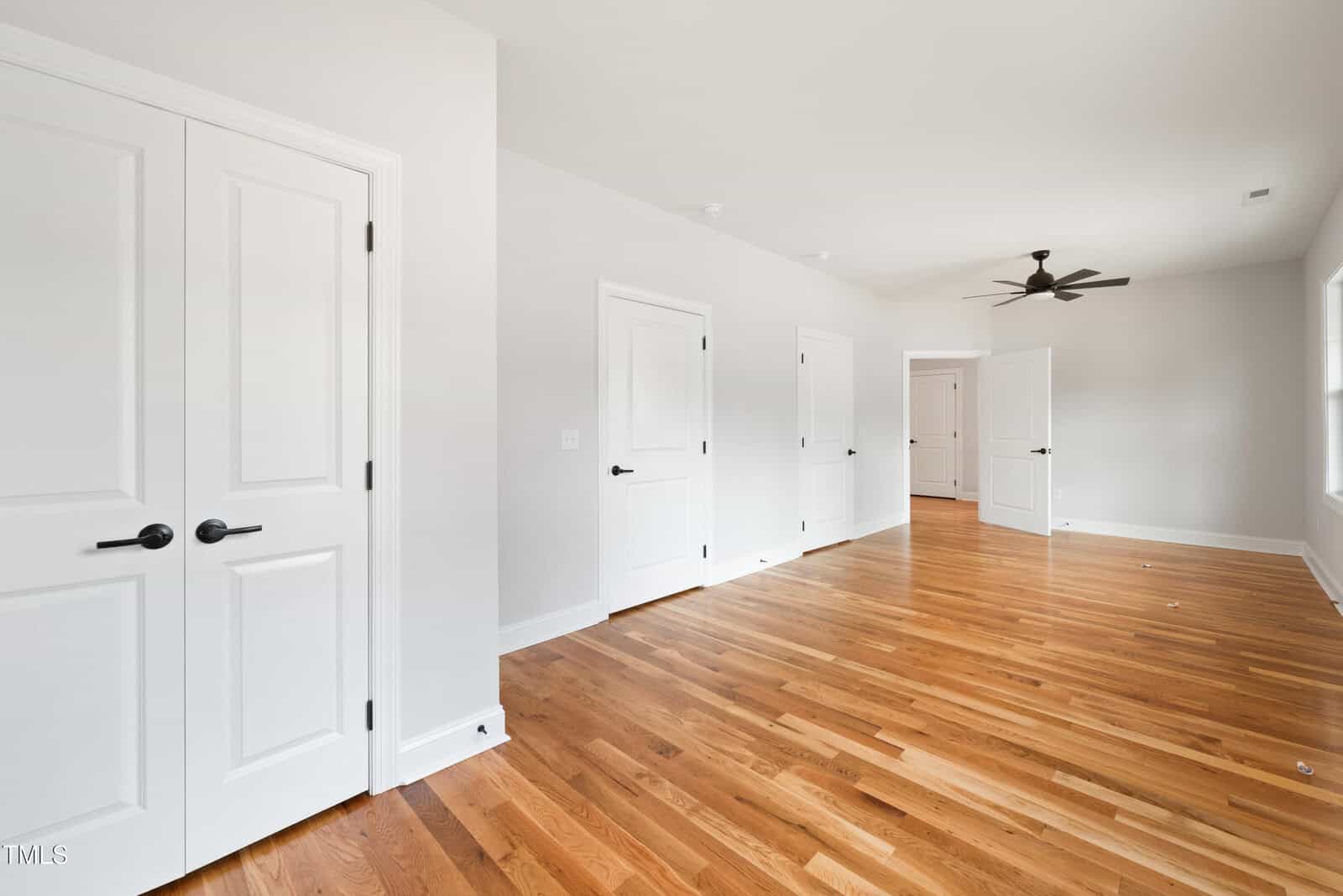Bright, empty room with light walls, hardwood floors, multiple white doors, a ceiling fan, and large windows letting in natural light.