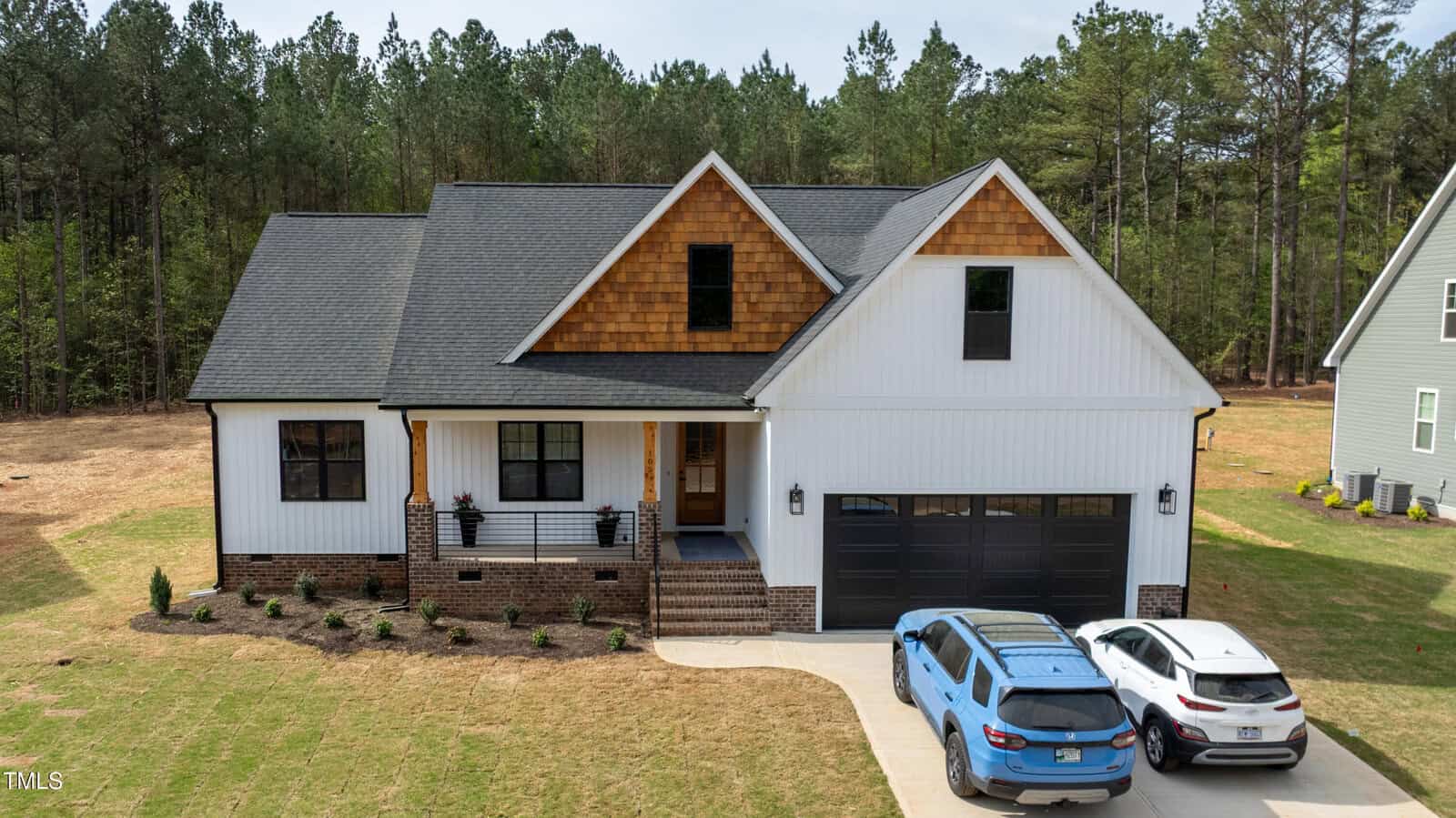 A two-story house with white siding, brown shingles on the upper front, a covered porch with columns, black garage doors, and two cars parked in the driveway. The yard has sparse grass and newly planted shrubs.