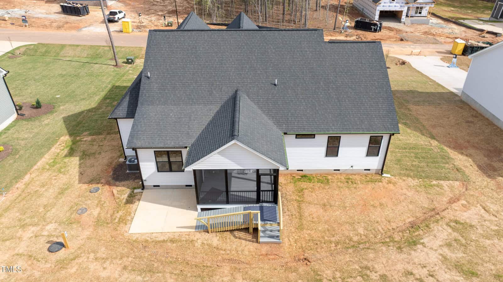 Aerial view of the back of a single-story house with a black roof, white siding, and a screened porch. The yard is mostly grass with some patches of bare soil. Nearby houses are visible under construction.