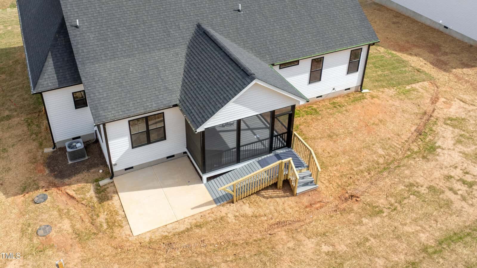 Aerial view of the back of a white house with a black roof, a screened porch, wooden steps with railings, a small concrete patio, and a patchy lawn.