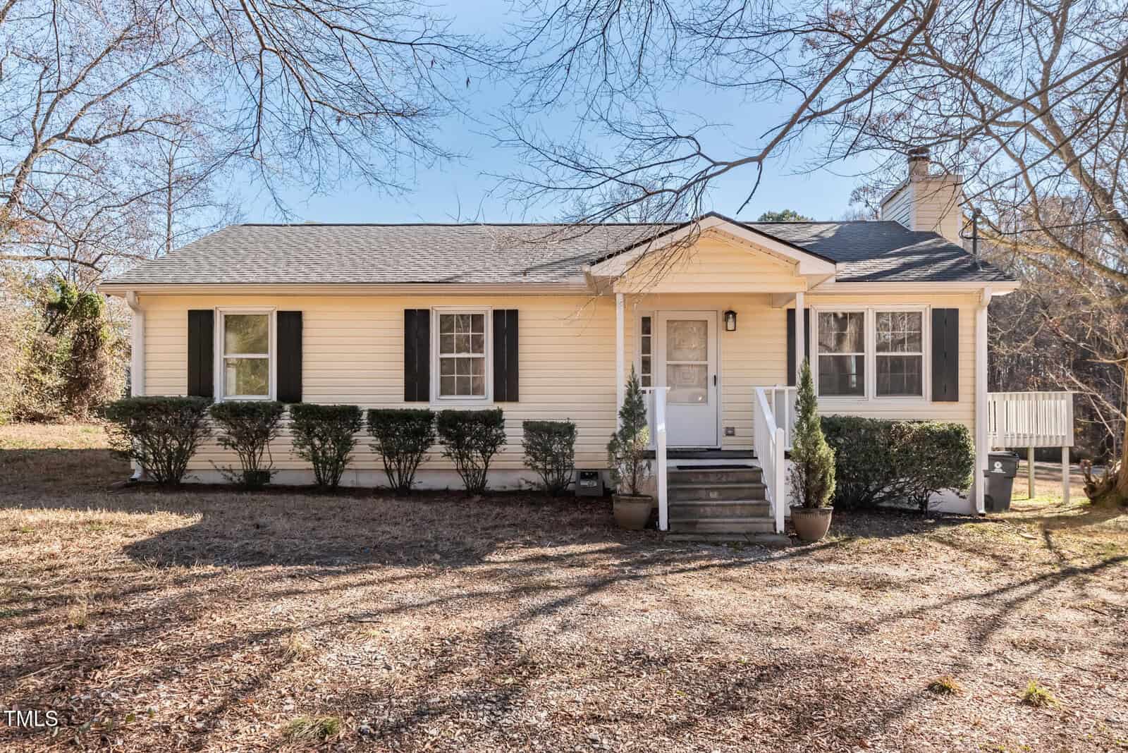 A single-story beige house with black shutters, a gray shingled roof, and a small front porch with steps. Leafless trees and patchy grass are visible in the yard on a clear day.