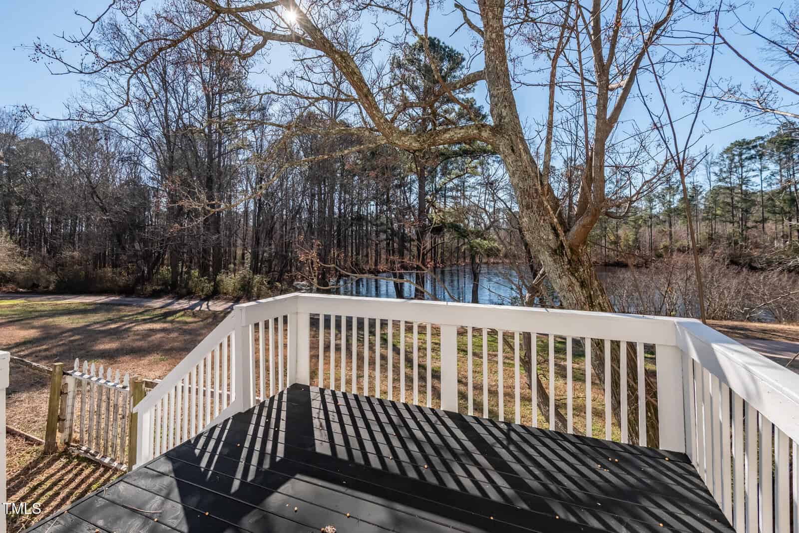 A white-railed deck overlooks a grassy yard with a wooden fence, trees, and a pond in the background under a clear blue sky.