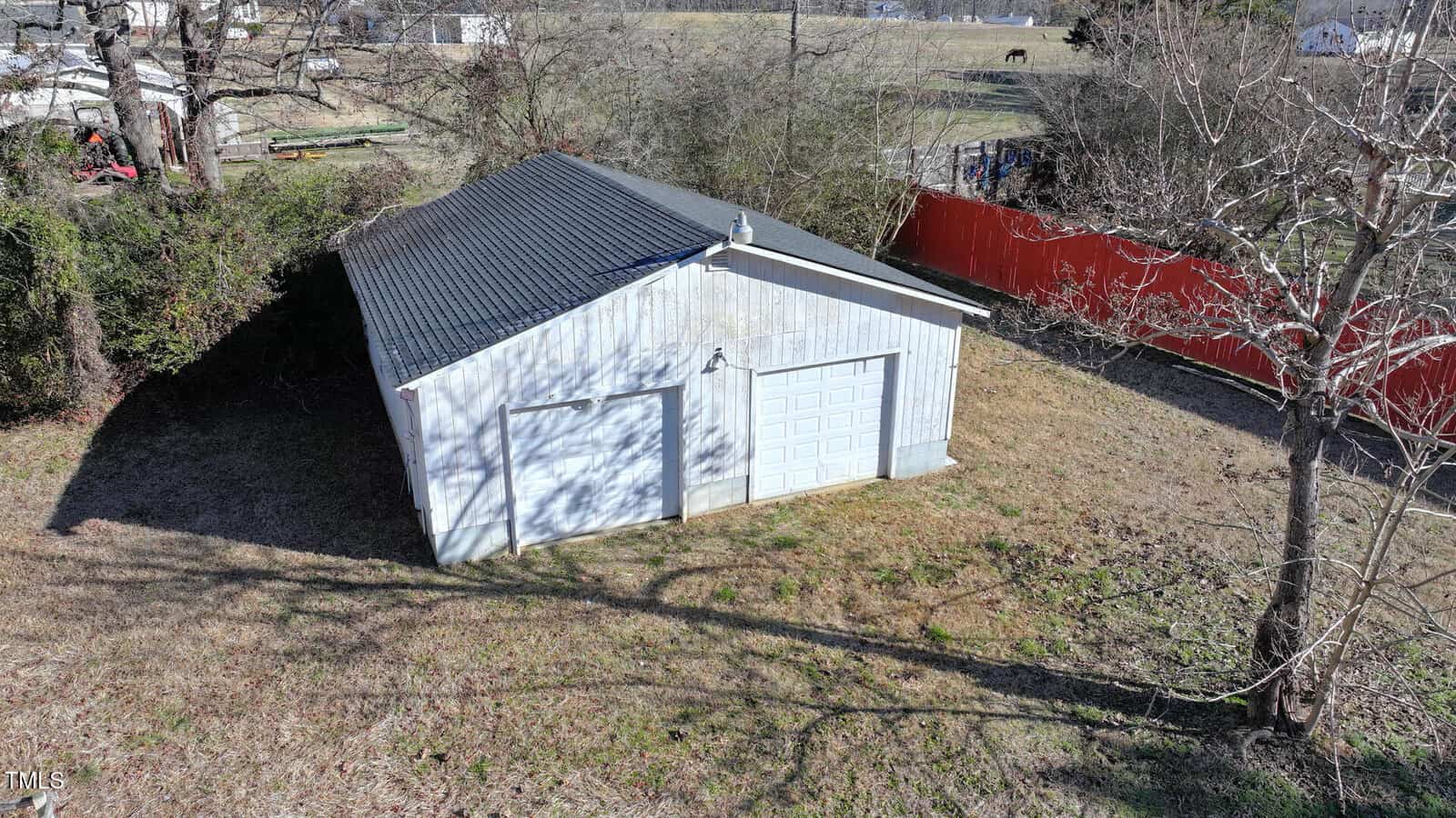 Aerial view of a white metal two-car garage with a gray roof, surrounded by grass and trees. A red fence and some distant buildings and fields are visible in the background.