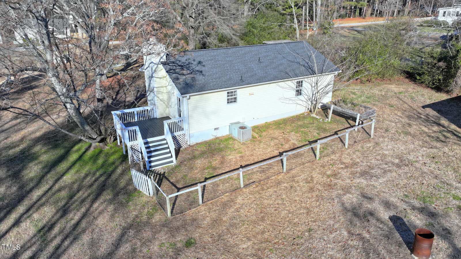 A small, light-colored house with a fenced backyard and steps leading to a raised deck, surrounded by leafless trees and dry grass in a rural area.