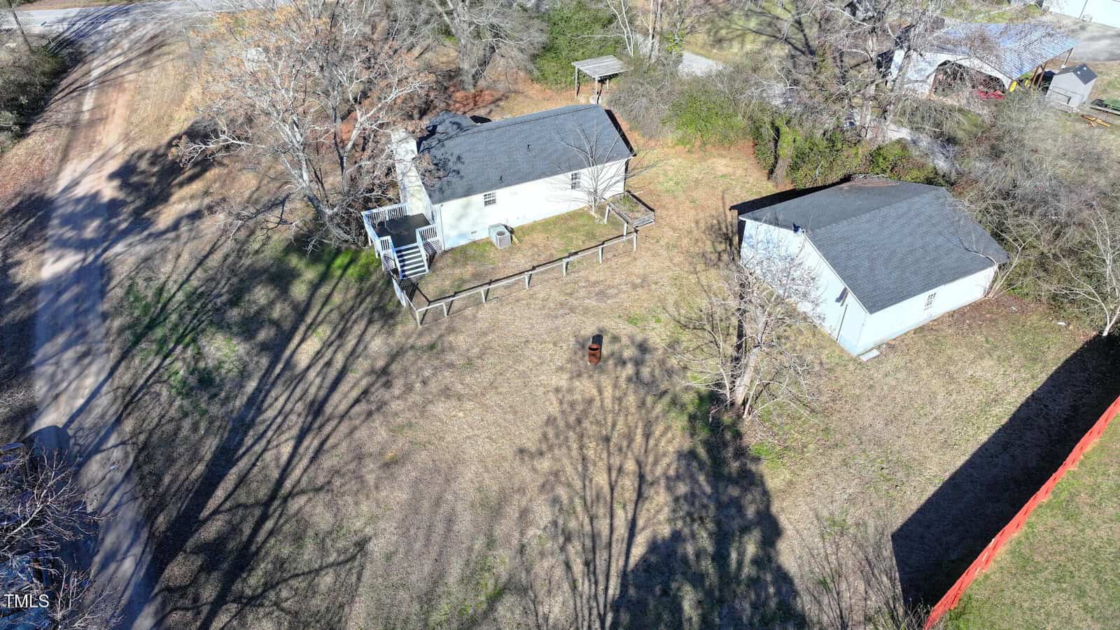 Aerial view of a small light-colored house with a deck and fenced yard, a detached garage or shed, leafless trees, and a dirt road on the left side. Shadows from the trees stretch across the grass.