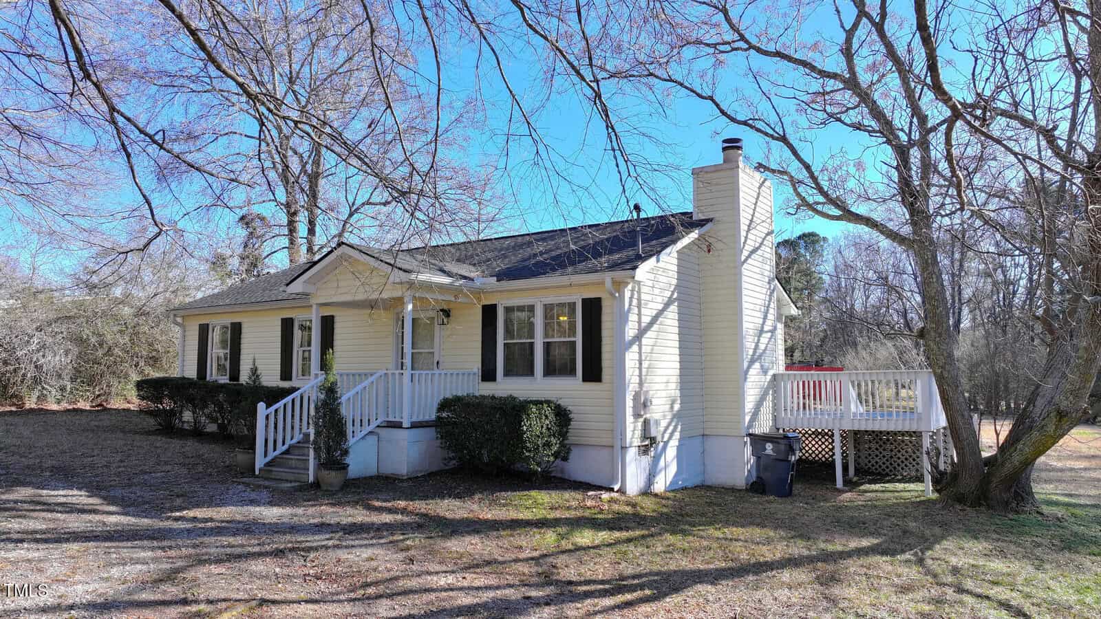 Small yellow house with white trim, front porch, and side deck. Surrounded by bare trees and shrubs, the house sits under a clear blue sky on a spacious, grassy lot.