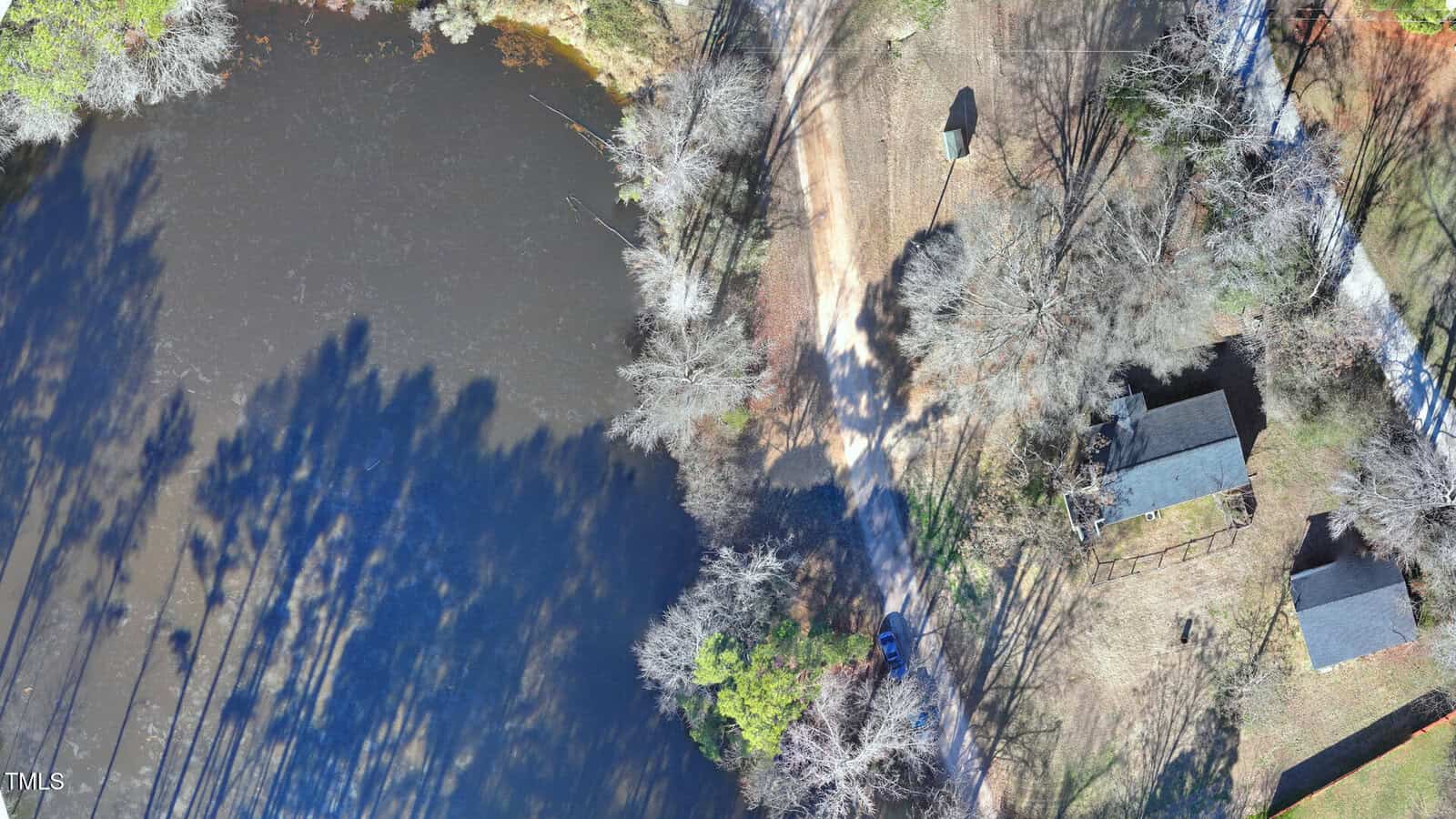 Aerial view of a small pond surrounded by leafless trees, a dirt road, a parked vehicle, and two adjacent dark-roofed buildings in a rural area on a sunny day.