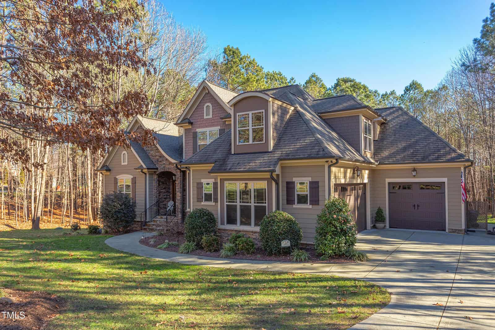 A beige two-story house with a stone facade, multiple gables, and a two-car garage. The driveway curves toward the entrance. It's surrounded by trees and has a well-maintained lawn.