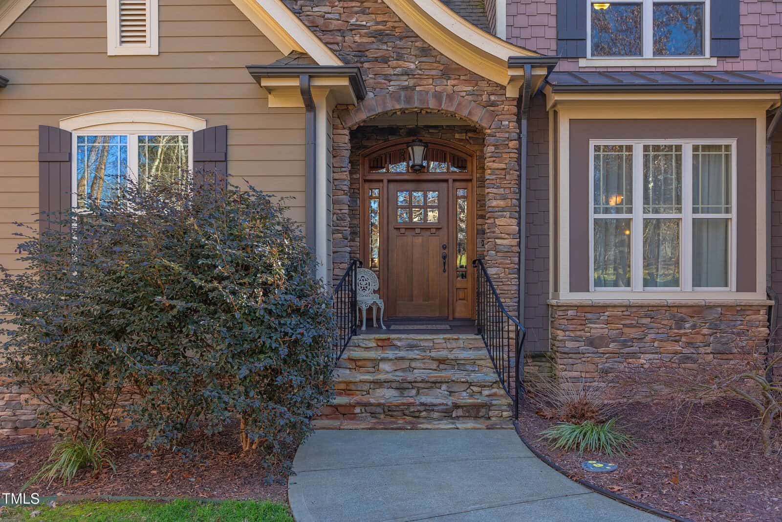Front entrance of a house with a stone facade and a wooden door. Steps lead up to the door, flanked by black metal railings. Bushes line the pathway, and there is a small decorative item on the steps. Two windows are visible on either side.