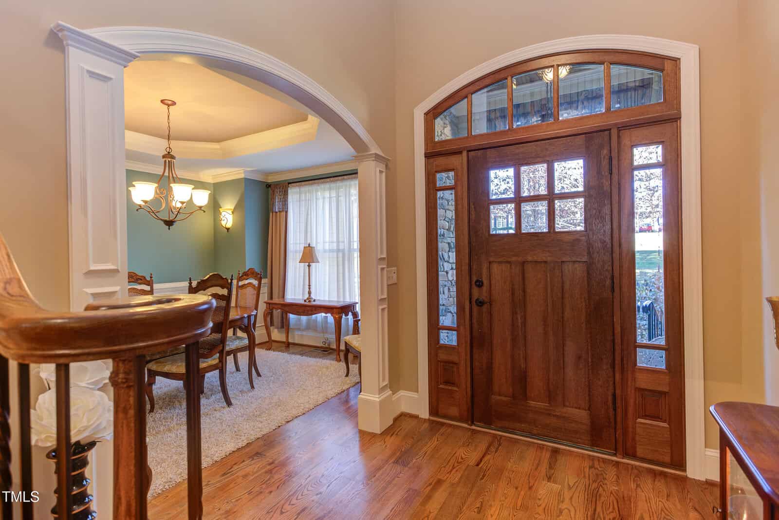 A foyer with a wooden front door featuring glass panels. Adjacent is an archway leading to a dining room with a chandelier, wooden chairs, and a round table. The spaces have hardwood floors and indoor lighting creates a warm ambiance.
