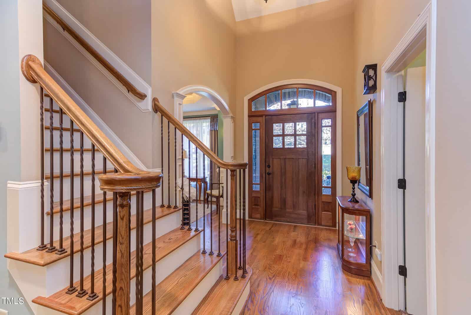 A brightly lit foyer with hardwood floors and a staircase with wooden handrails and white risers. A wooden front door with glass panels is at the center. Arched doorways lead to adjacent rooms, and a small table is against one wall.