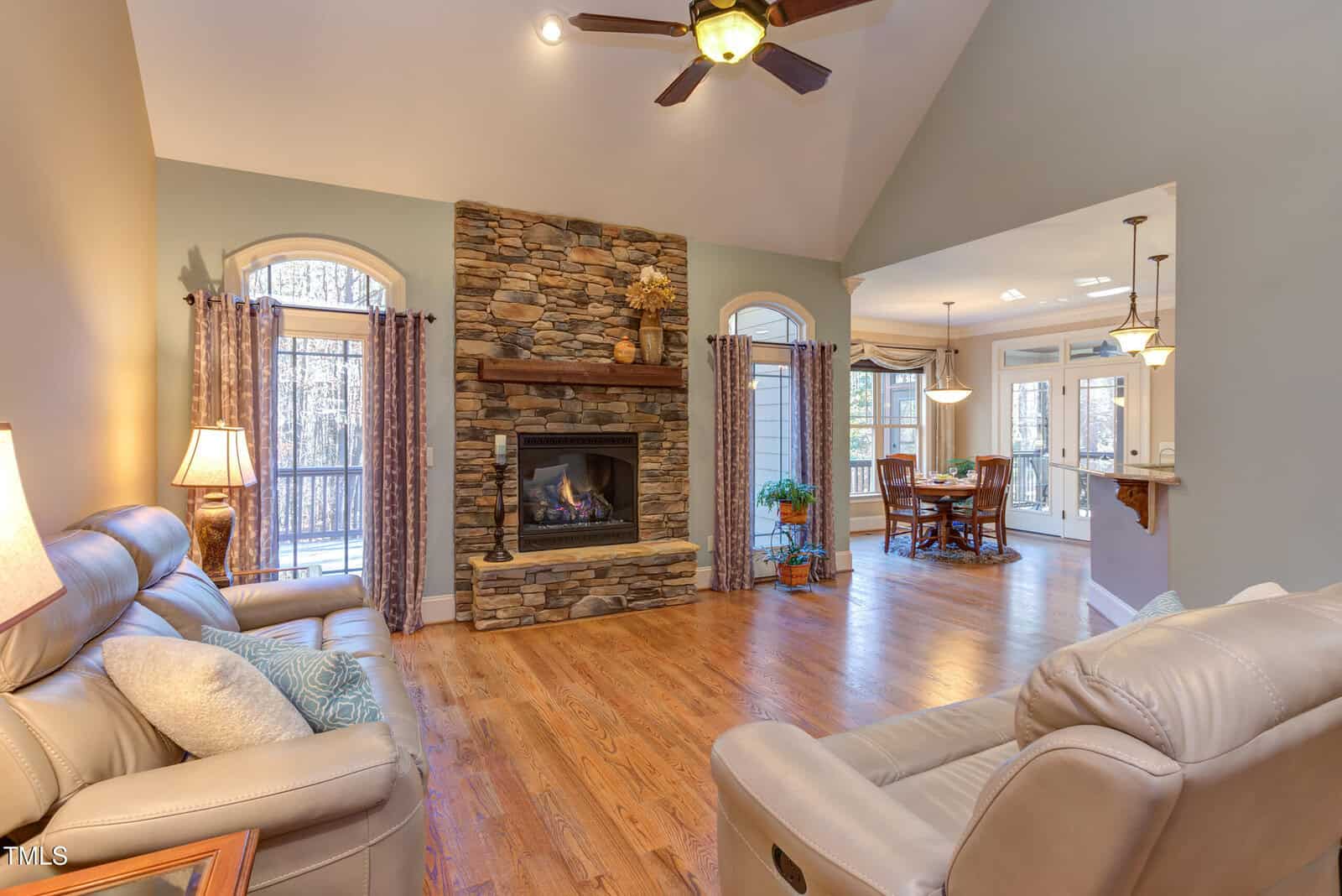 Living room with a stone fireplace, leather sofas, and wooden floor. Ceiling fan above. Opens to a dining area with a table and chairs. Large windows allowing natural light. Neutral colors and decorative lamps are present.