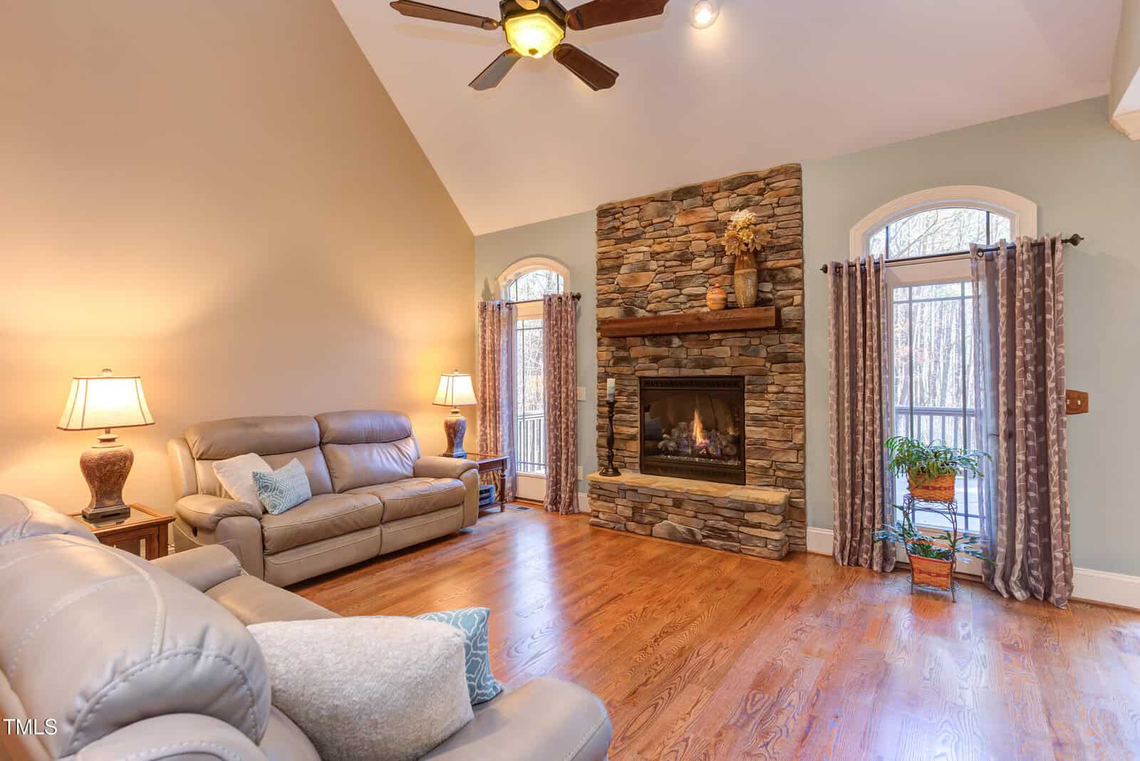A cozy living room with high ceilings features two gray leather sofas and a stone fireplace. Natural light enters through two arched windows with curtains. A wooden floor and a ceiling fan complete the room's warm and inviting atmosphere.