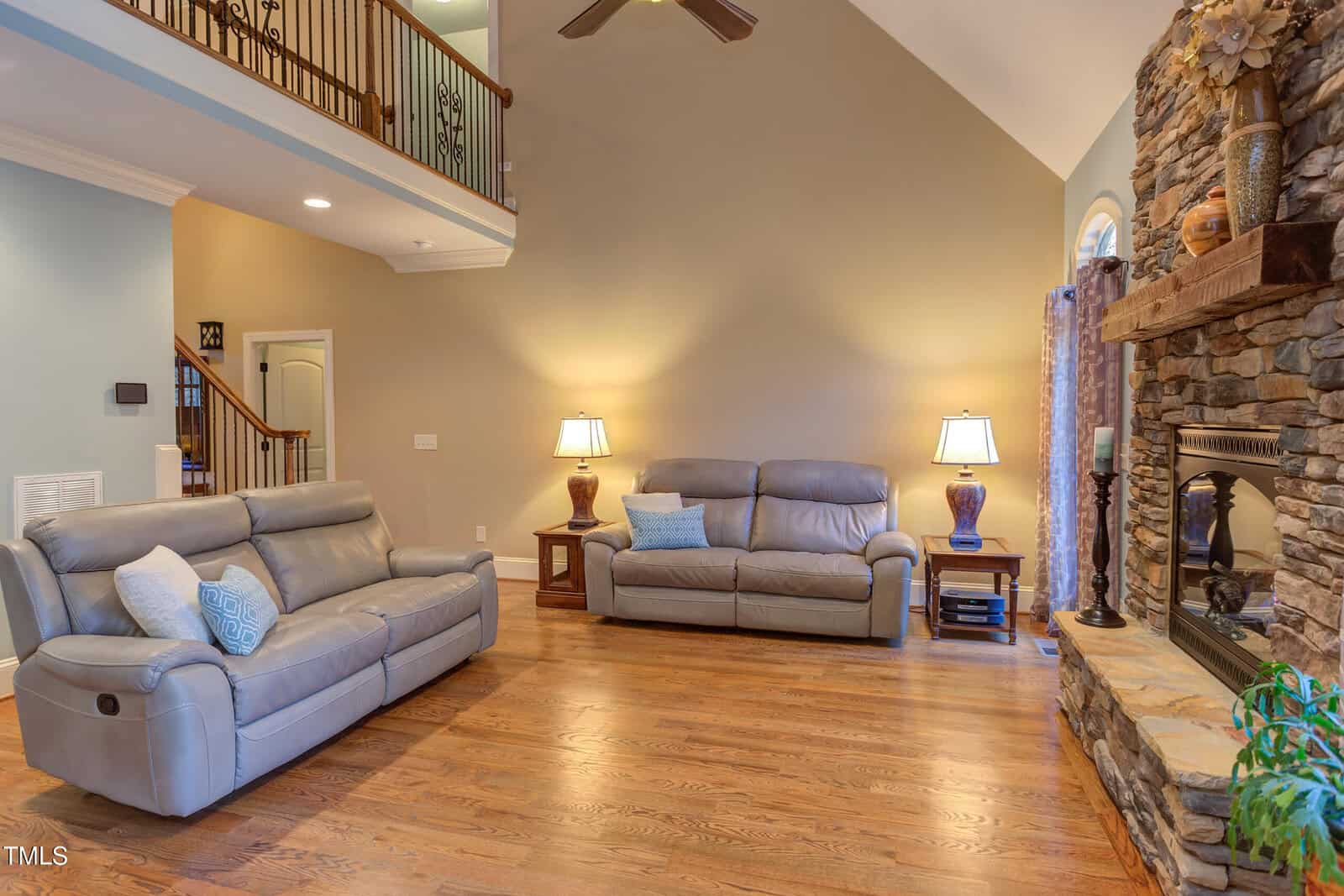 A spacious living room with high ceilings features two gray leather sofas, a stone fireplace, and wooden flooring. Two lamps stand on side tables next to the sofas. A staircase with a wooden banister leads to the upper floor.