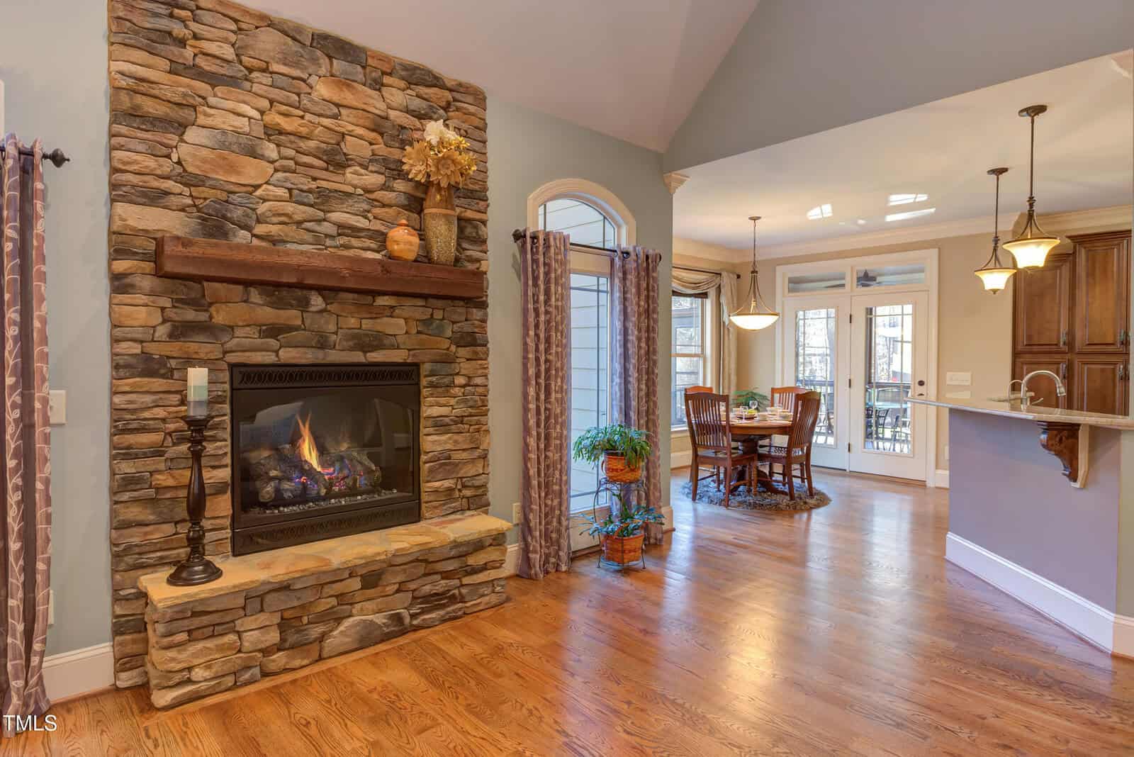 A cozy living space with a stone fireplace on the left, featuring a wooden mantel. The room has hardwood floors and an adjacent dining area with a table set for four. The area is well-lit with natural light and hanging pendant lamps.