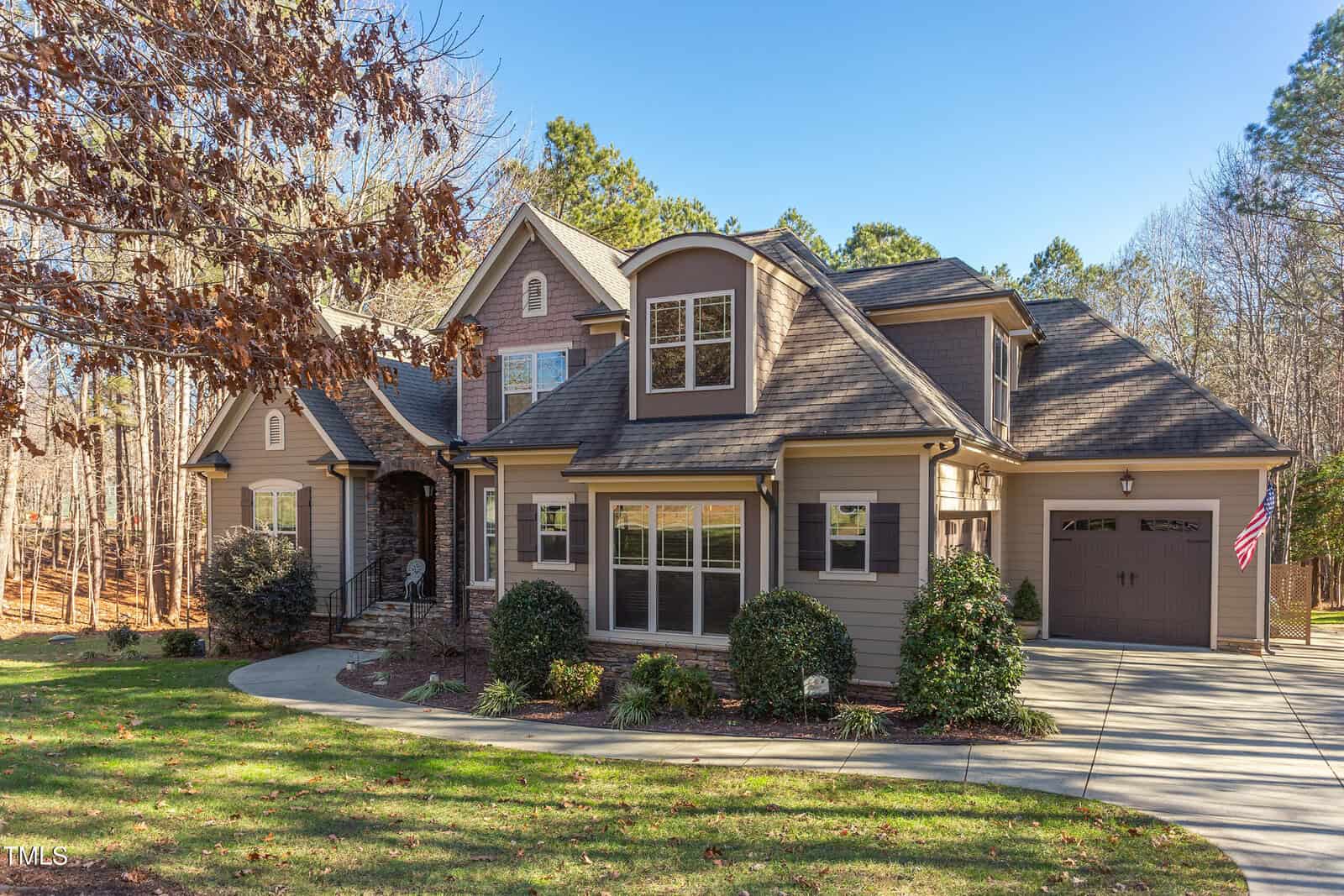 Located at 512 Belmont Circle in Wake Forest, this two-story house boasts a brown and gray exterior with a gabled roof and two-car garage. The curved driveway is framed by a well-maintained lawn and shrubs, while trees stand tall in the background under a clear blue sky.