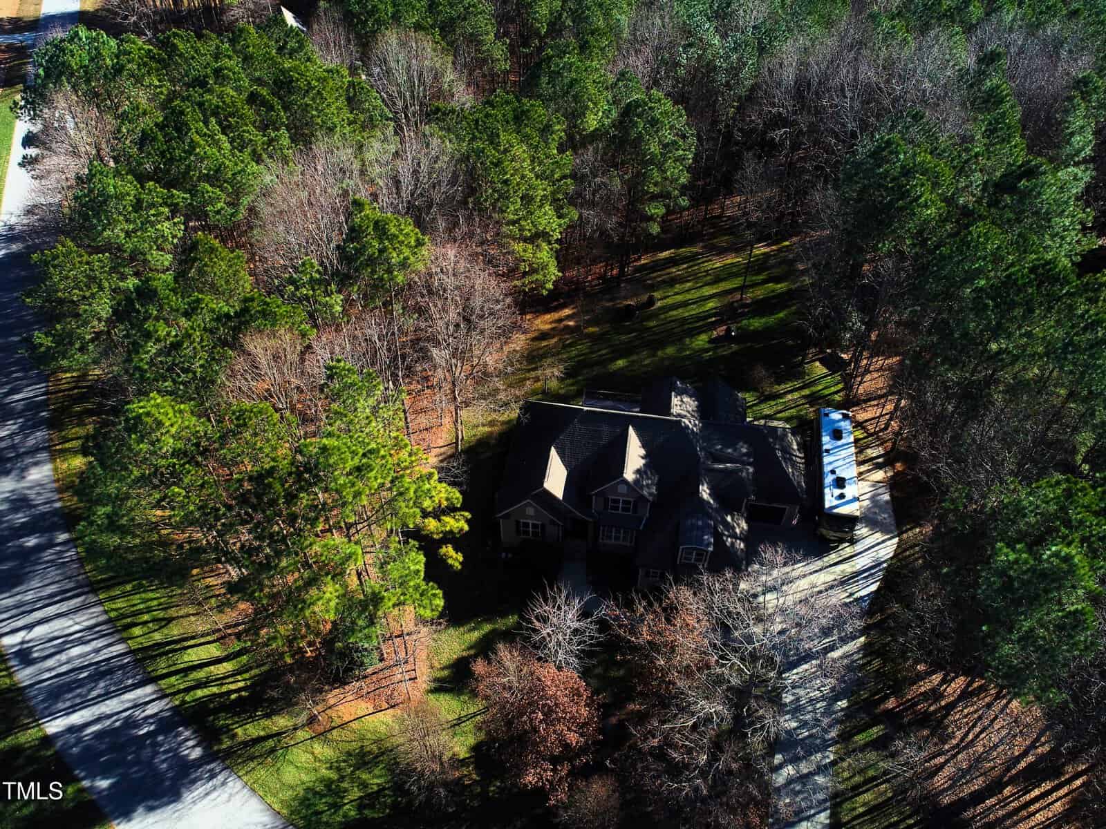 Aerial view of a large house surrounded by a dense forest of trees. The property includes an RV parked on the driveway and a winding road in the bottom left corner. Dense greenery encircles the home, blending with the surrounding landscape.