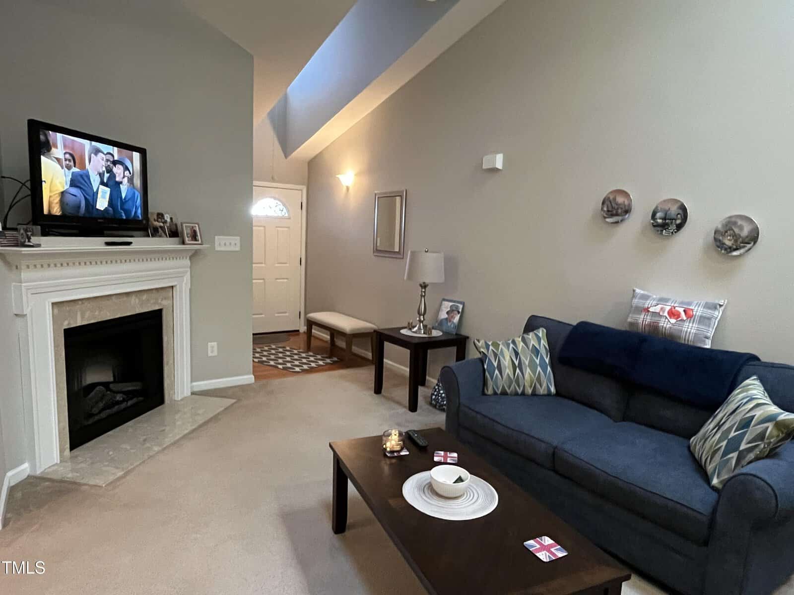 A living room with beige walls and carpet, featuring a dark blue couch, a wooden coffee table, and a fireplace with a TV above it. Decorative plates hang on the wall, and a lamp sits on a side table next to the couch.