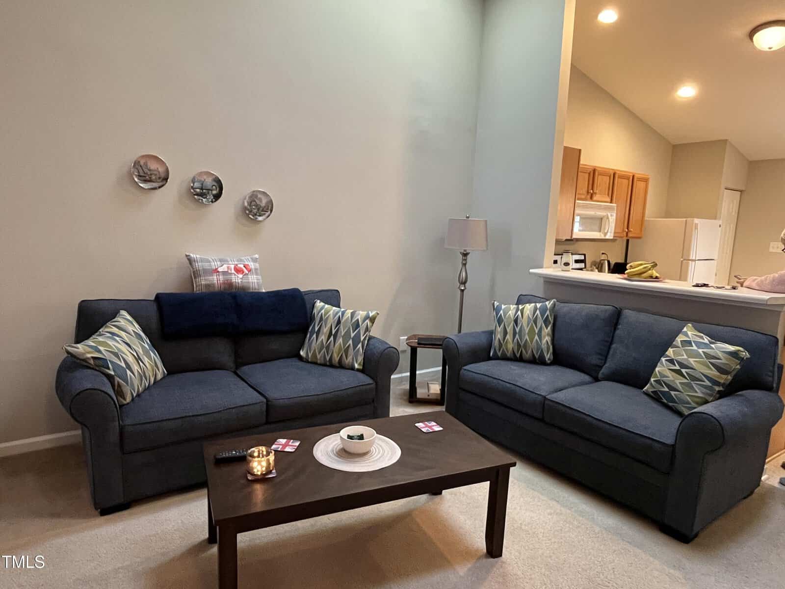 A living room with two blue sofas adorned with patterned pillows, a wooden coffee table with coasters, a candle, and a bowl. The background shows a kitchen area with light-colored cabinets and a counter. A floor lamp stands between the sofas.