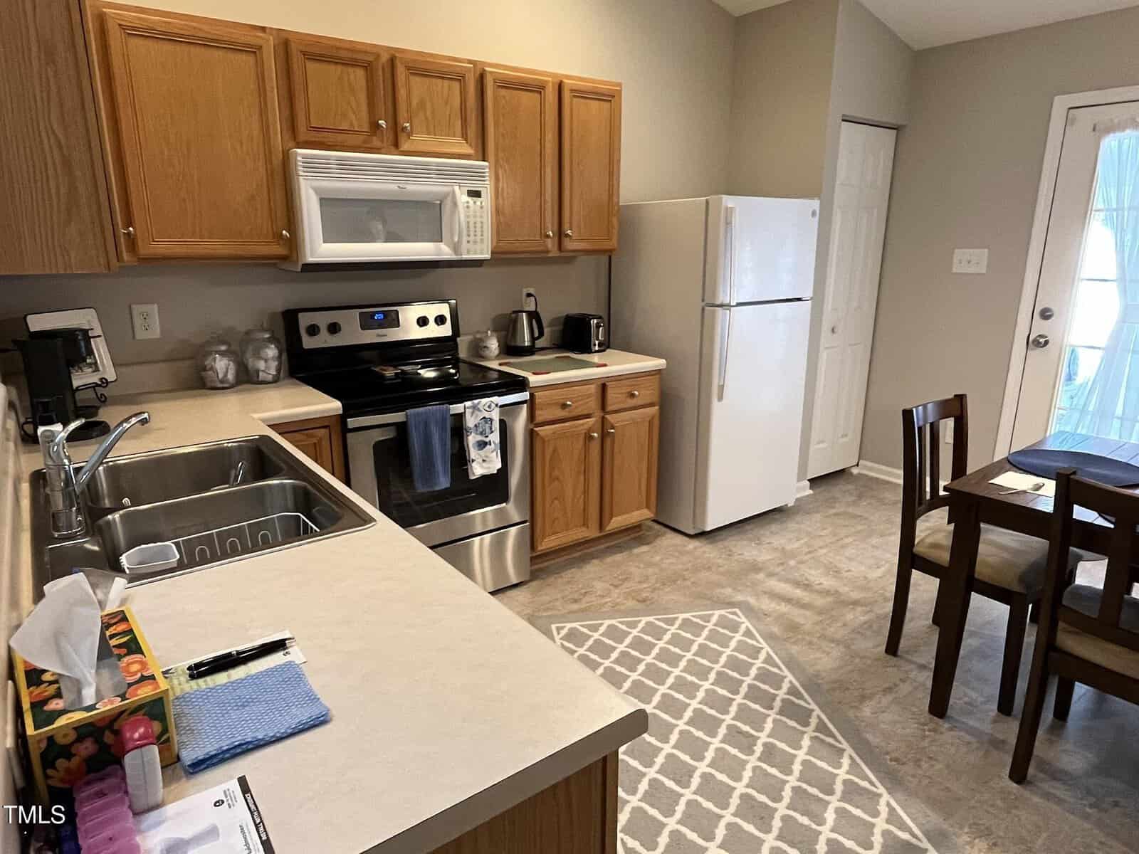 A kitchen with light brown cabinets, a white refrigerator, and stainless steel oven and microwave. There's a double sink, countertop appliances, and a small dining table with chairs. A rug sits on the floor, and a door leads outside.