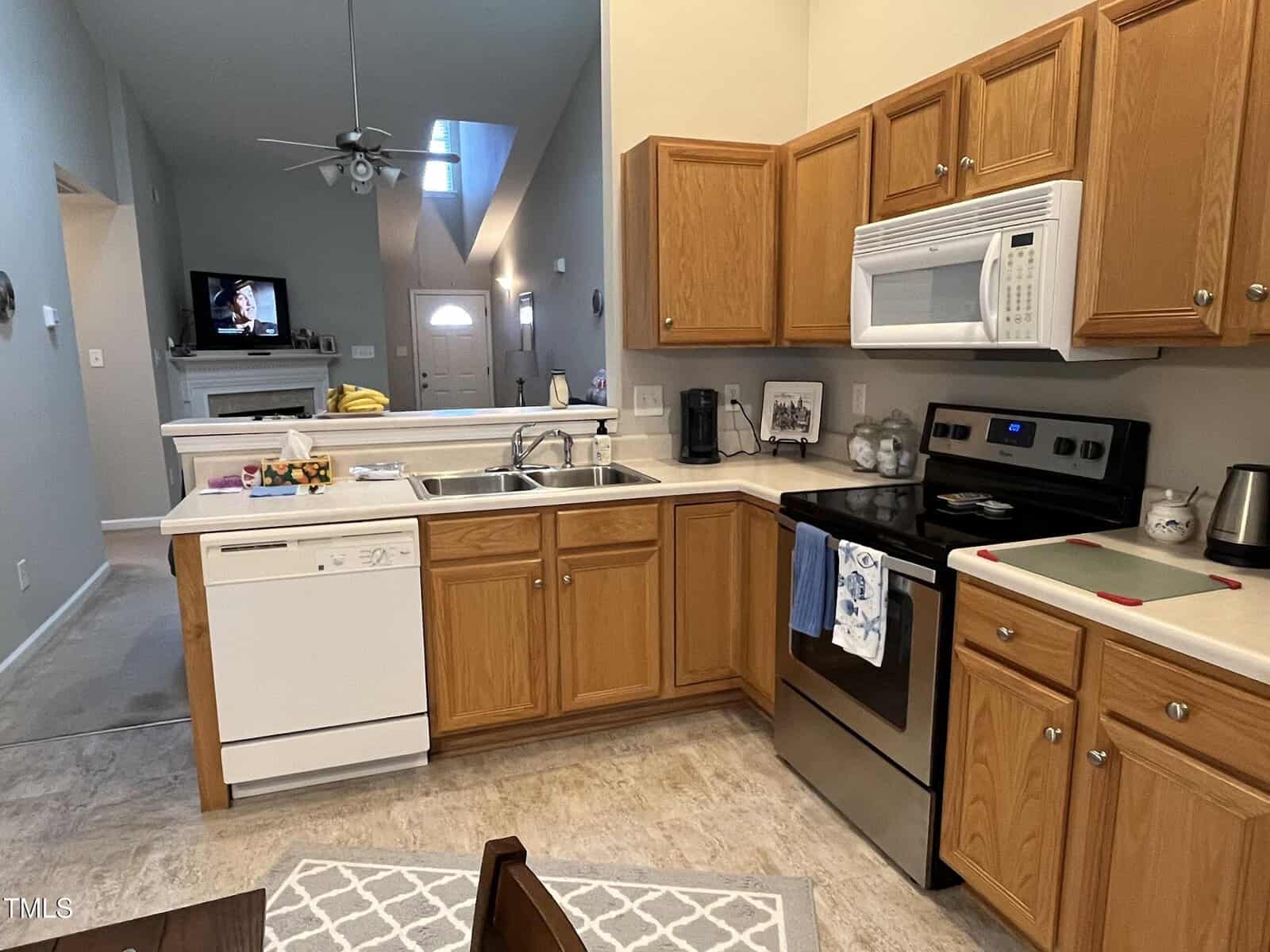 A kitchen with wooden cabinets, a stainless steel stove, and a white dishwasher. A microwave is mounted above the stove. The sink is under a window. A small dining table is partially visible. A living area with a TV and ceiling fan is in the background.