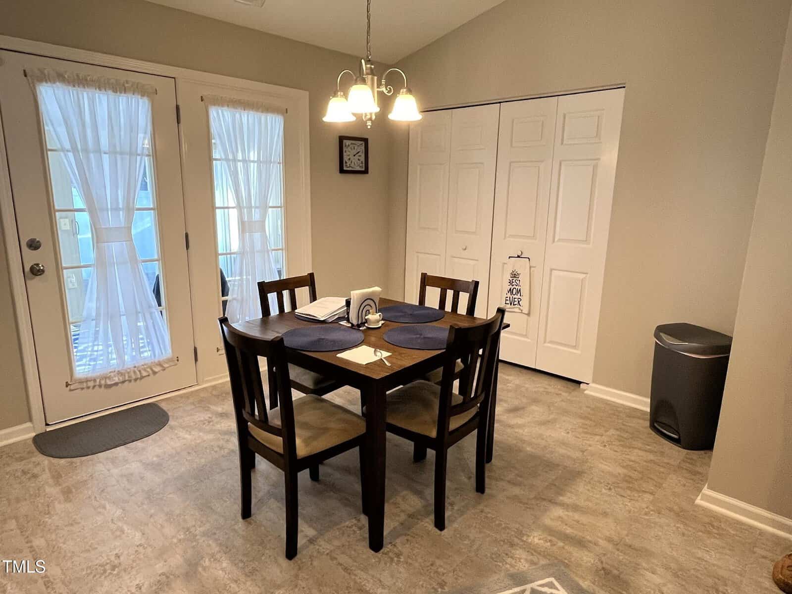 A dining area features a wooden table with four chairs, set with blue placemats. A light fixture hangs above. There are double doors with sheer curtains on the left and a white closet with a decorative door hanger on the right. A trash bin is nearby.