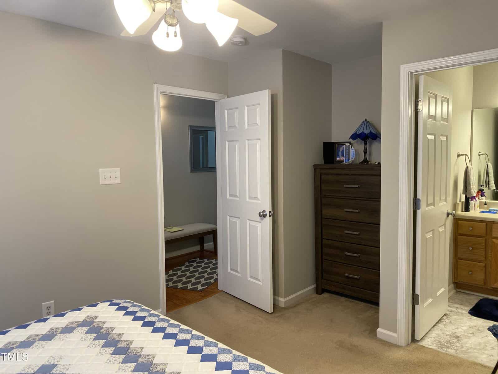 A bedroom with a ceiling fan and a bed with a blue and white checkered quilt. There is a wooden dresser with a blue lamp, a door leading to a bathroom, and another door leading to a hallway with a bench and rug visible.
