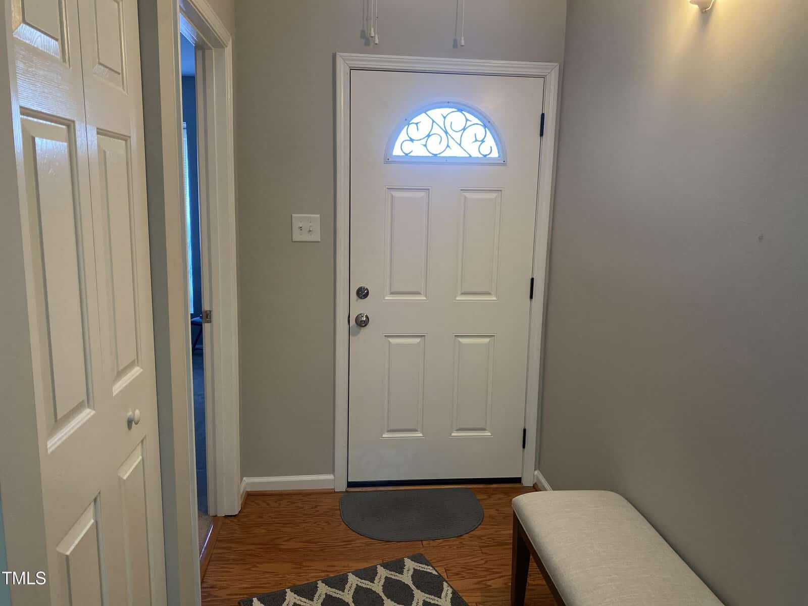 A small entryway with a closed white door featuring an ornamental window at the top. The floor is wooden with a gray rug and a beige bench on the right. The walls are painted light gray, and there are coat hooks above the door.