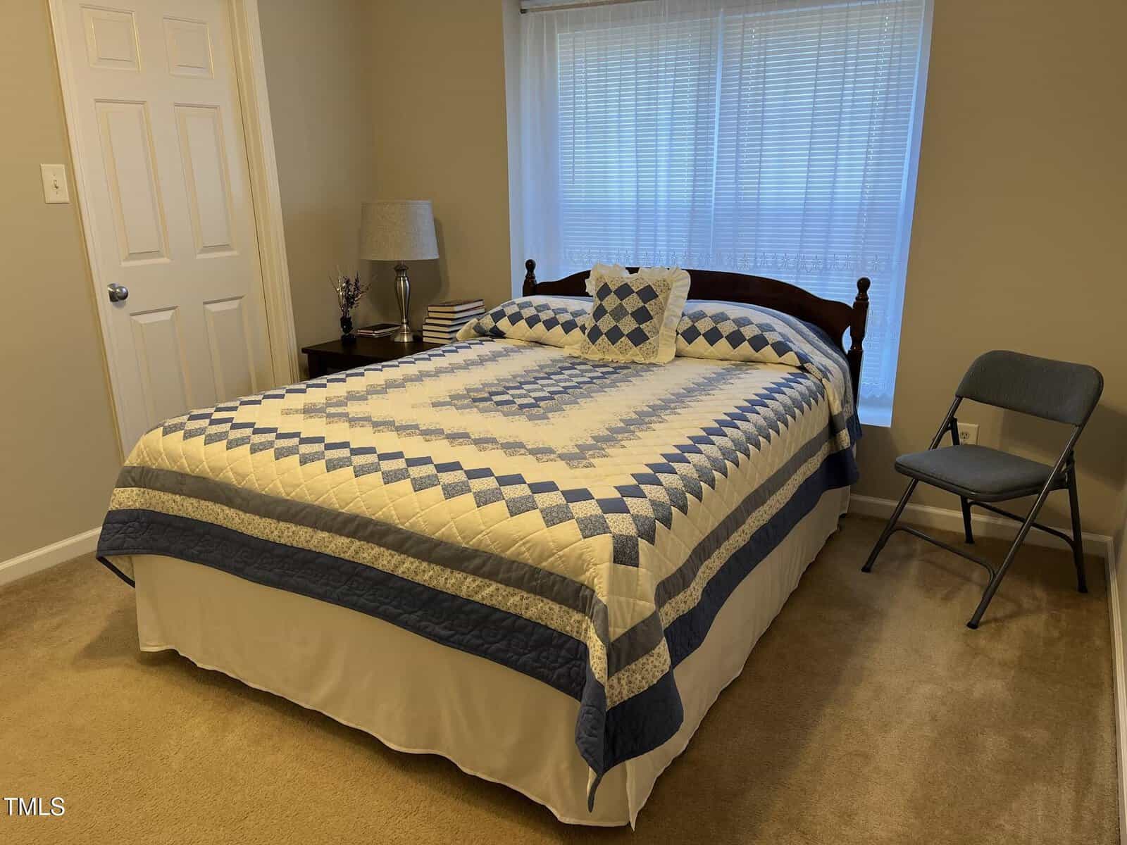 A bedroom with a neatly made bed featuring a blue and white checkered quilt, a headboard, and a decorative pillow. A small lamp and books sit on the nightstand. A folded chair is placed near the window with closed blinds. Carpeted floor visible.
