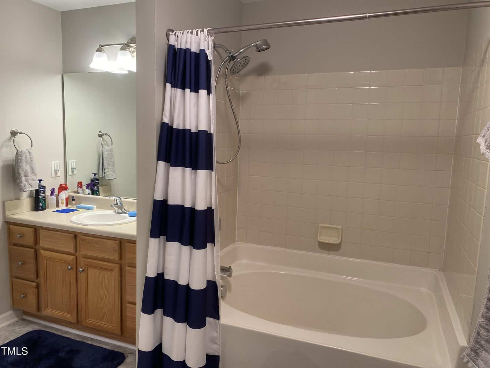 Bathroom with a combined bathtub and shower, featuring white tiles and a blue and white striped curtain. To the left, there is a sink with a wooden cabinet, a mirror, and various toiletries.