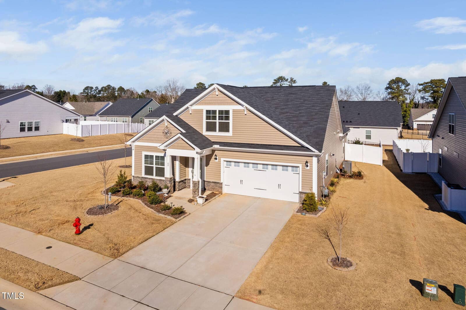 A beige, single-story house with a two-car garage and a gable roof sits on a corner lot with a well-maintained lawn, minimal landscaping, and a white fenced backyard under a partly cloudy sky.