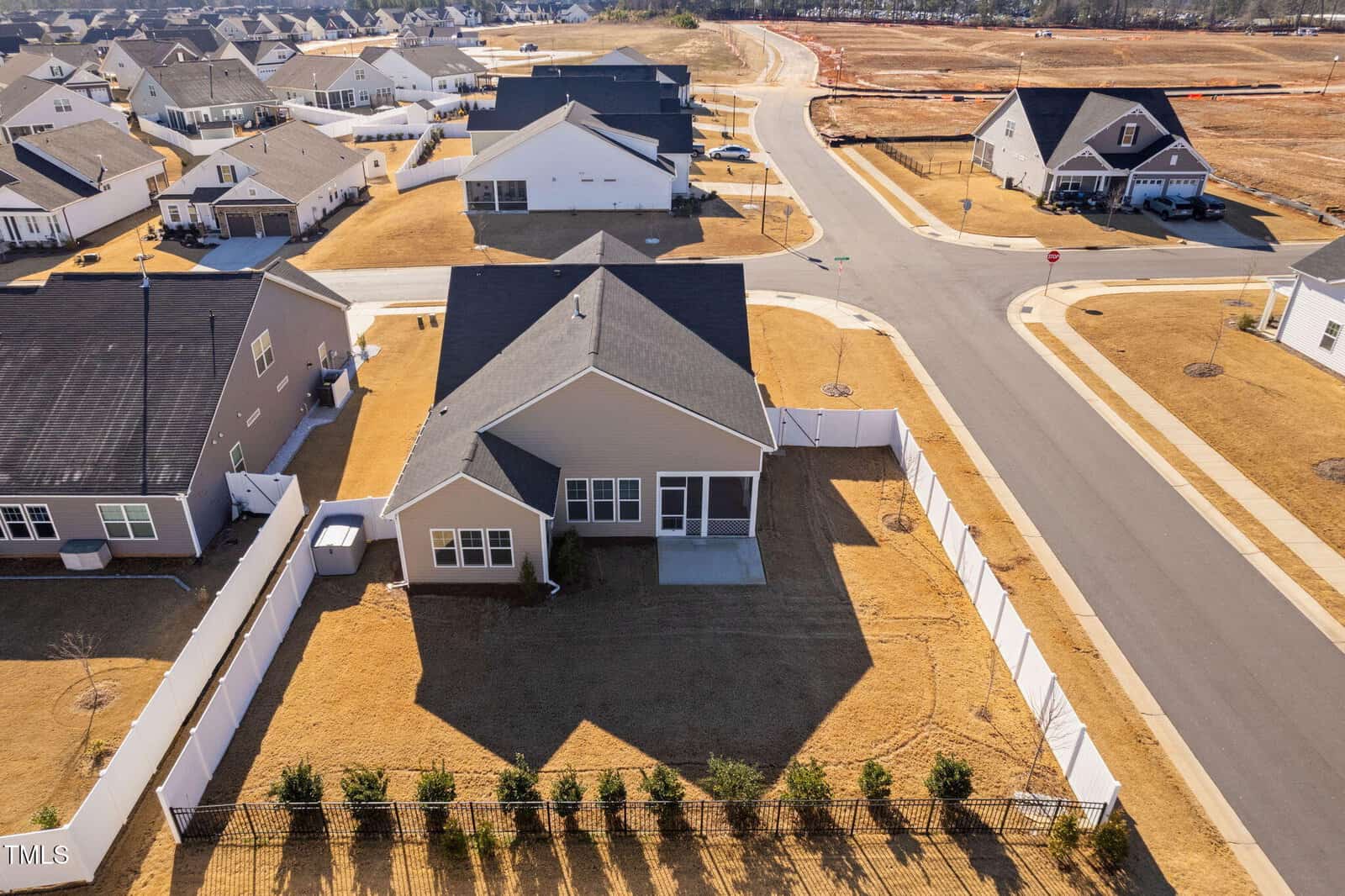 Aerial view of a single-story house with a fenced backyard, concrete patio, and surrounding homes in a residential neighborhood. Streets and sidewalks border the property, and the lawn is dry and yellow.