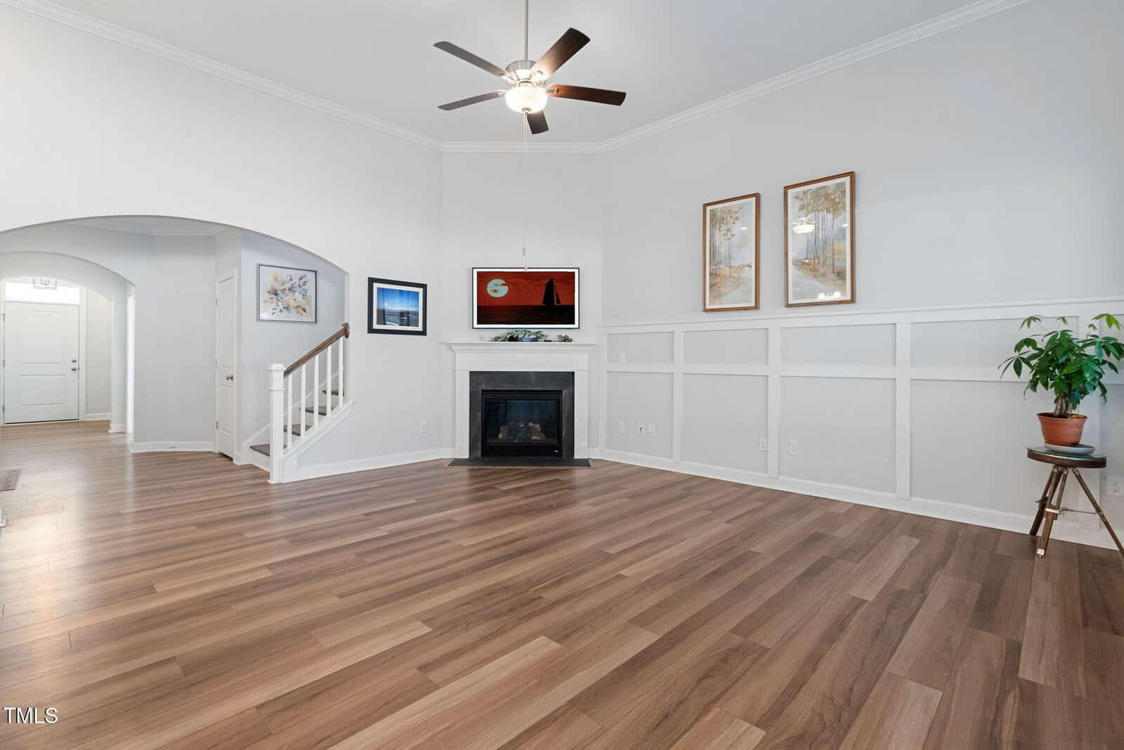 A living room with light wood flooring, white wainscoting, a corner fireplace, three framed pictures on the wall, a potted plant, a ceiling fan, and view of a hallway and stairs.
