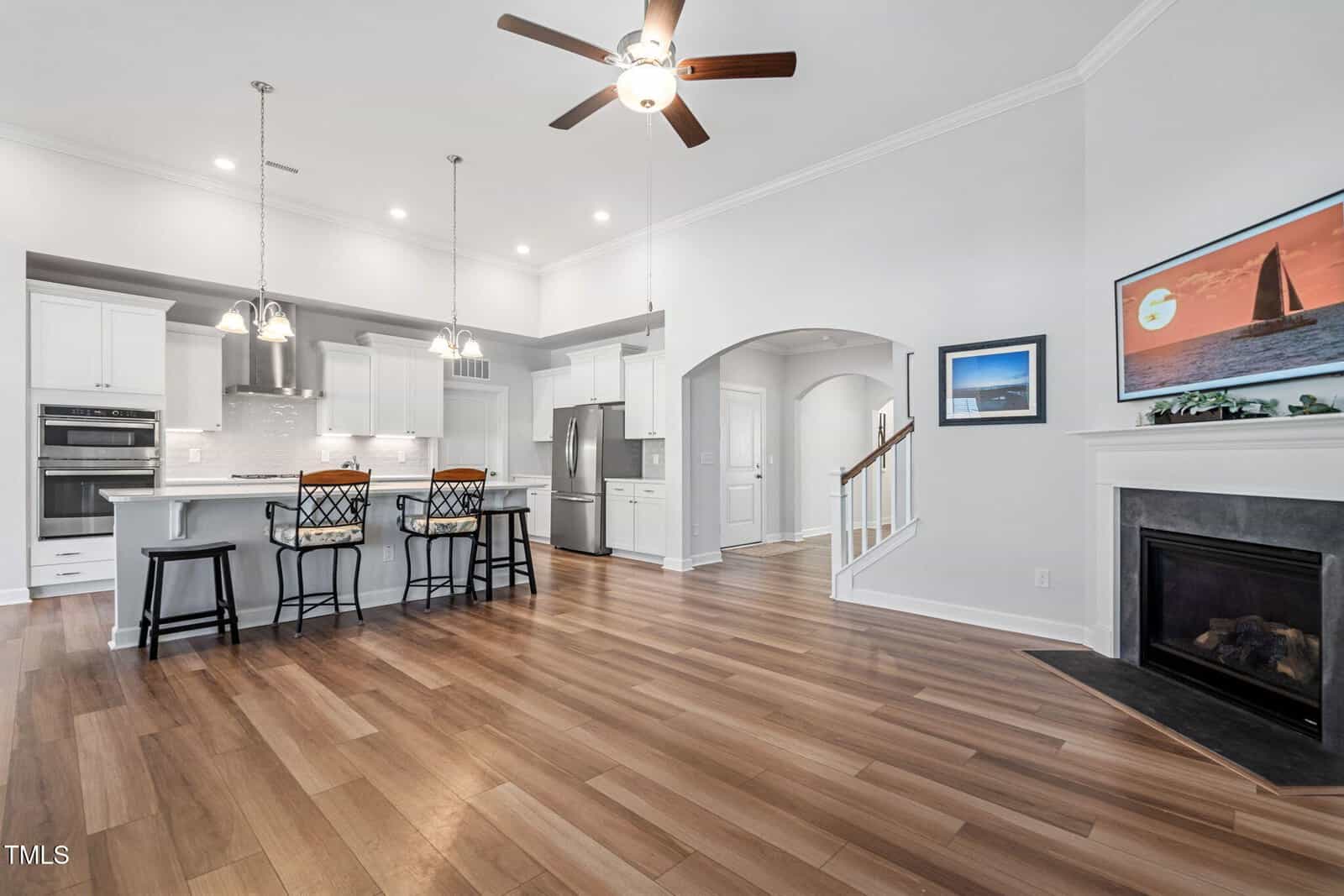 Modern kitchen and living area with wood floors, white cabinets, stainless steel appliances, a ceiling fan, island with barstools, fireplace, and framed art on the wall. Stairs lead up from the living space.