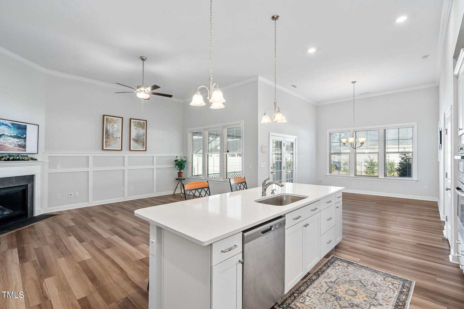 Open-concept kitchen and living area with white cabinets, island with sink, stainless steel appliances, pendant lights, ceiling fans, large windows, wood flooring, and a small rug near the sink.