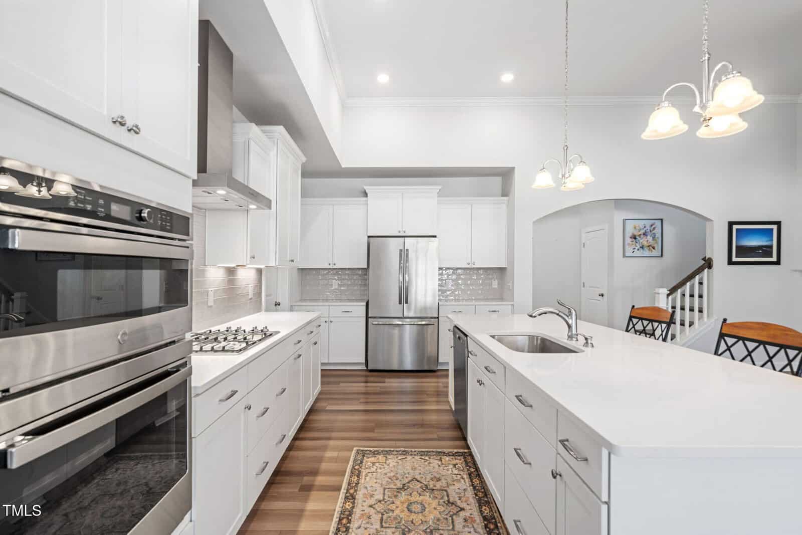 Modern kitchen with white cabinets, stainless steel appliances, a gas cooktop, double ovens, a large island with sink, pendant lights, wood flooring, a rug, and a staircase in the background.