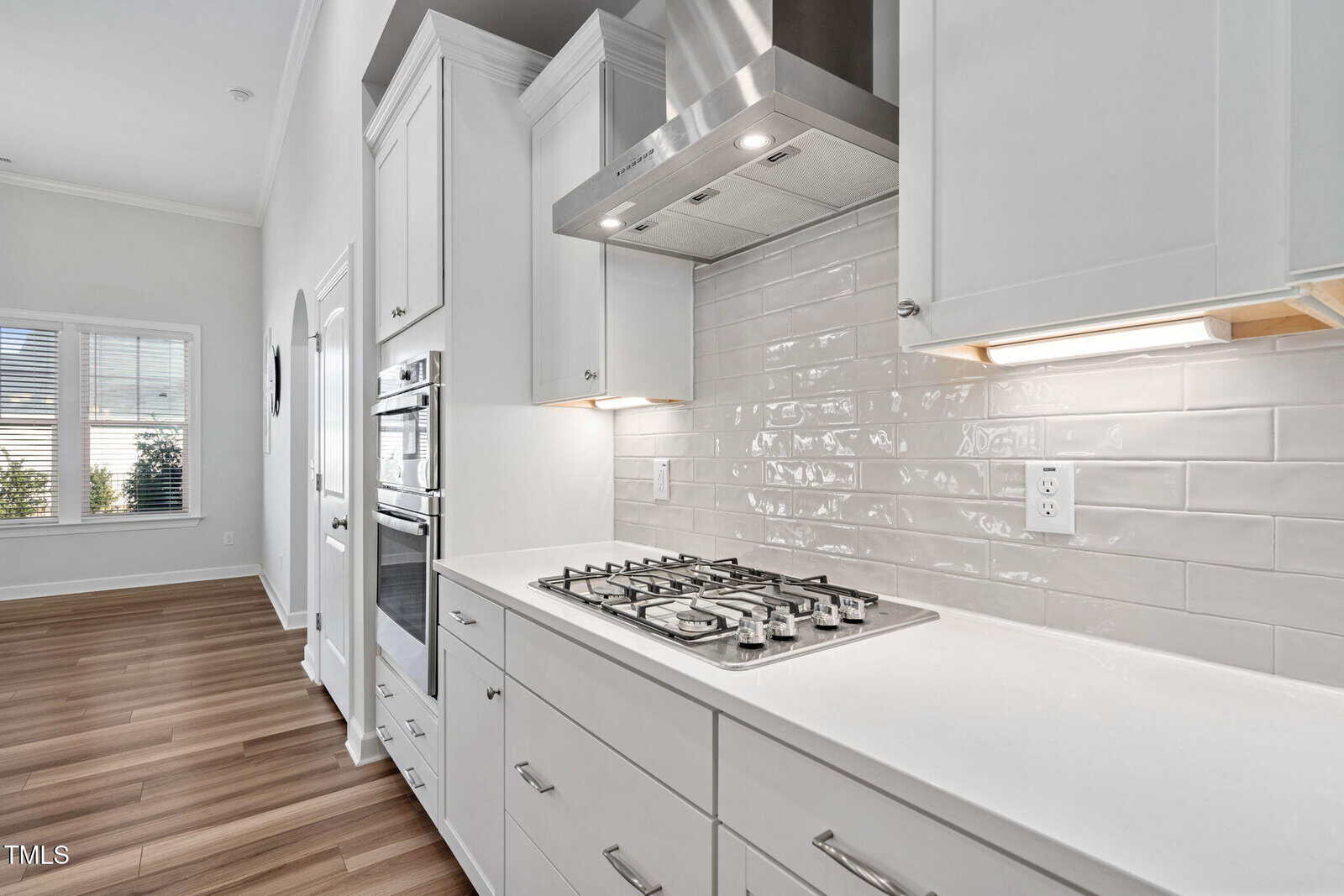 Modern kitchen with white cabinets, stainless steel range hood, built-in double ovens, and a gas cooktop. The backsplash is white subway tile, and the floor is medium-toned wood. Large window and arched doorway are visible.