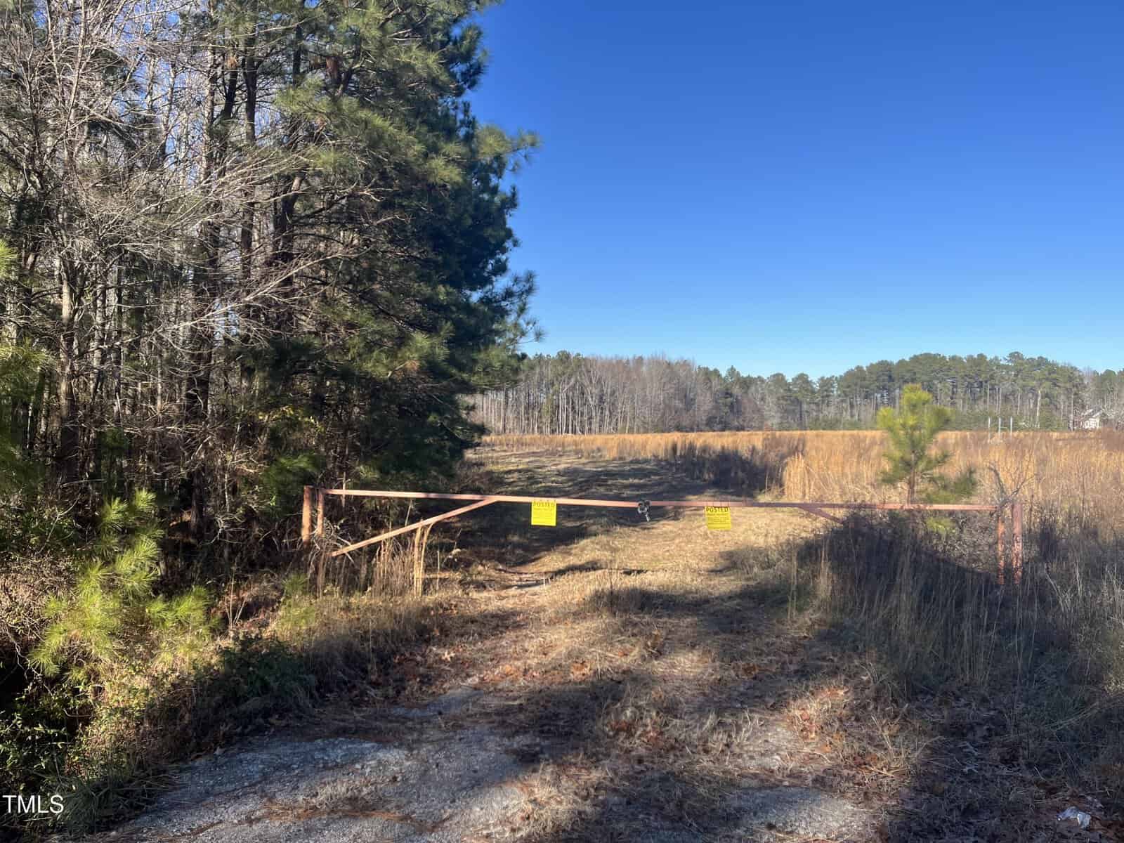 A metal gate blocks a gravel path leading to an open field with tall grass. Trees line the left side of the path, and a clear blue sky is visible above. Yellow warning signs are attached to the gate.