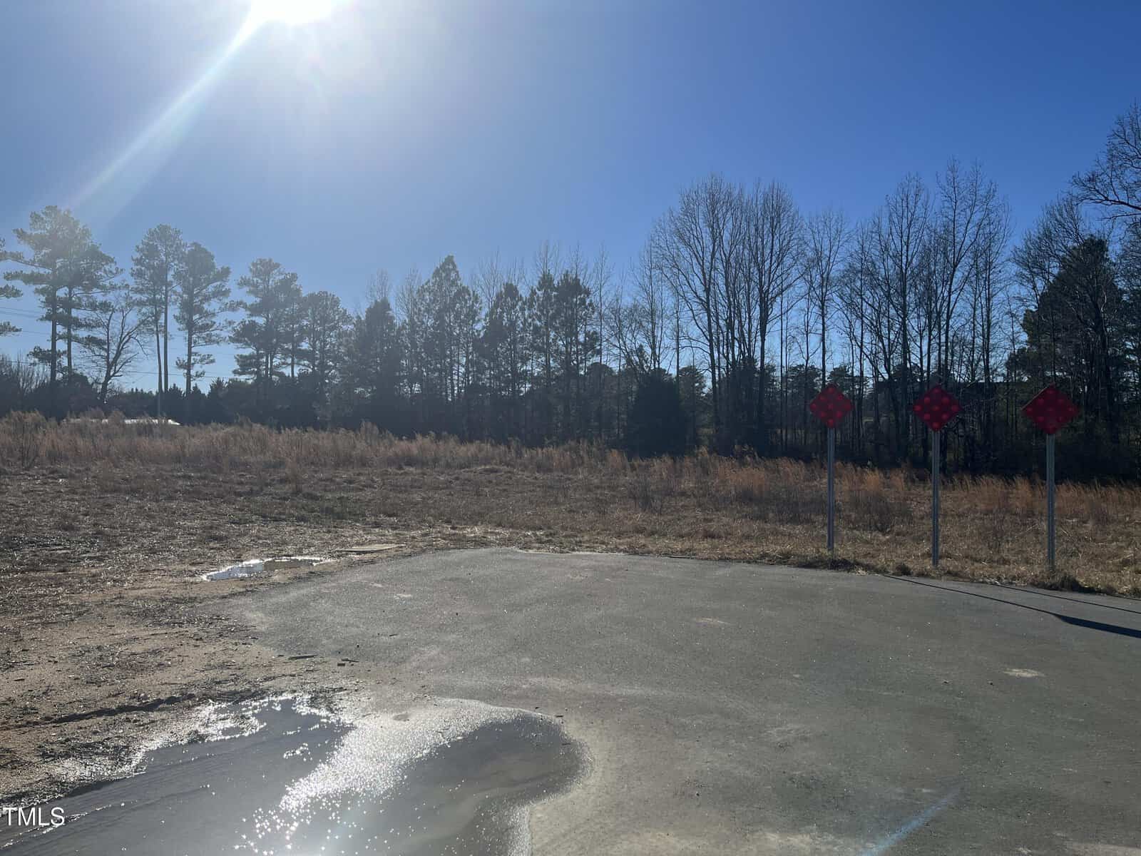 A paved road ends at a cul-de-sac with three dead-end signs. Grass, weeds, and trees are visible beyond the road under a bright blue sky with the sun shining.