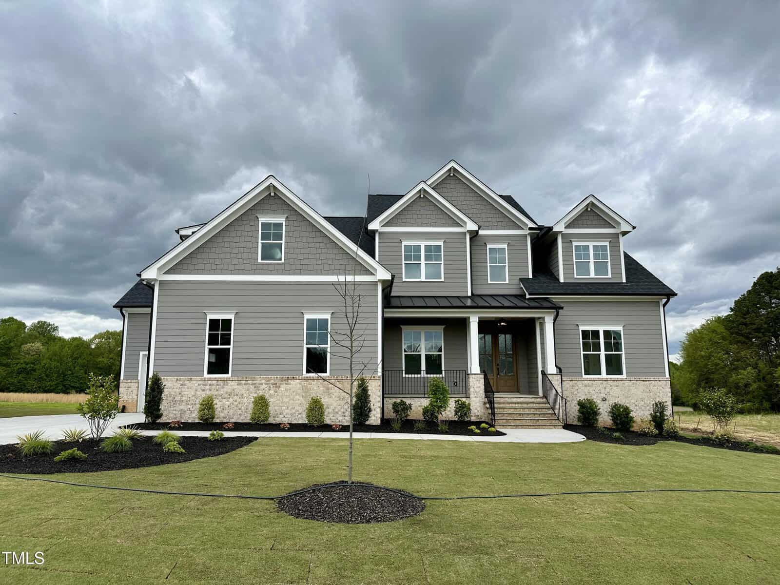 A two-story house with gray siding, brick accents, and a covered front porch sits under a cloudy sky. The front yard is landscaped with mulch, small shrubs, and a young tree.