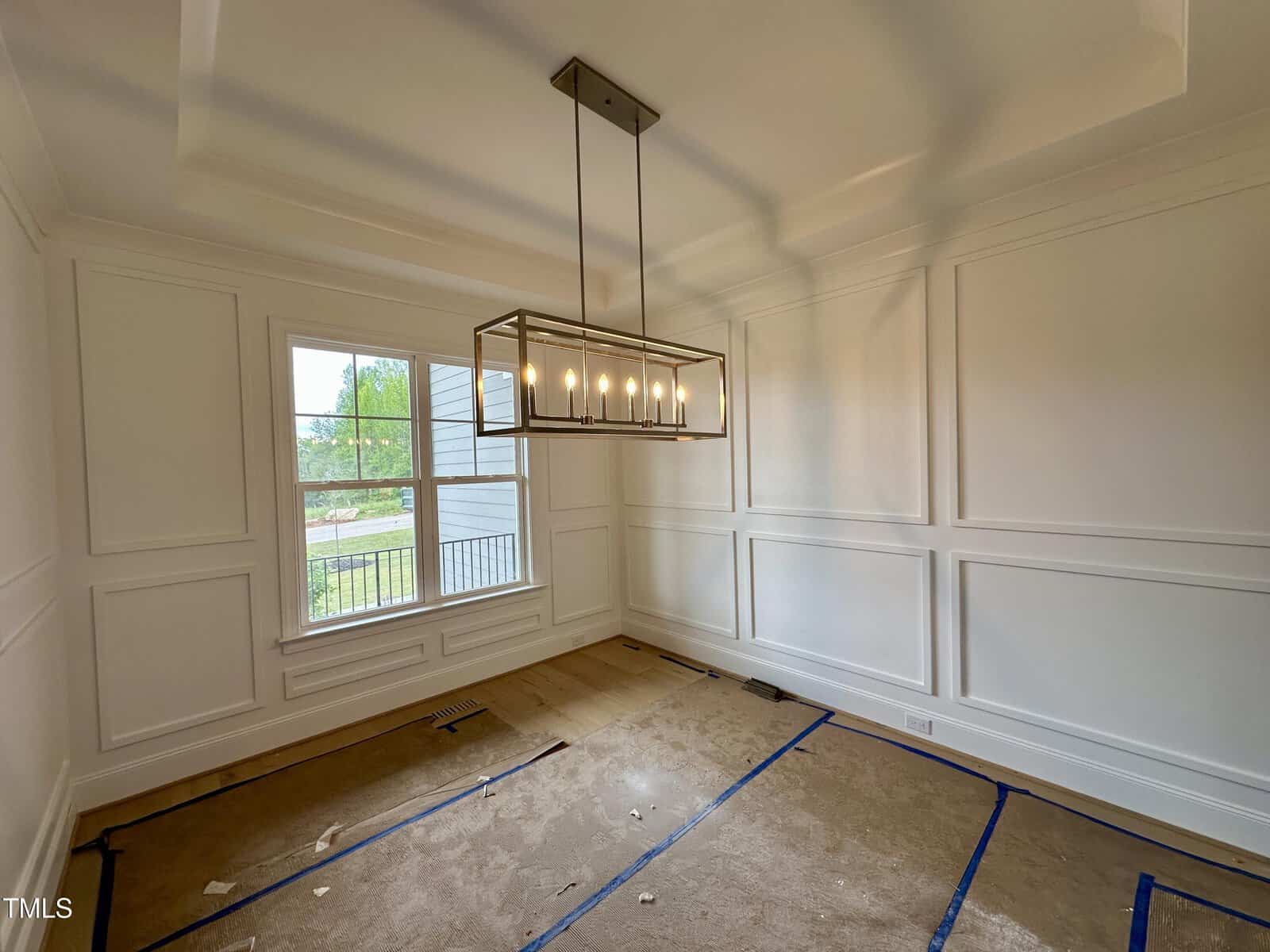 A mostly empty room with paneled white walls, a large window, a modern rectangular chandelier, and floors covered in protective cardboard and blue tape. Light comes in through the window.