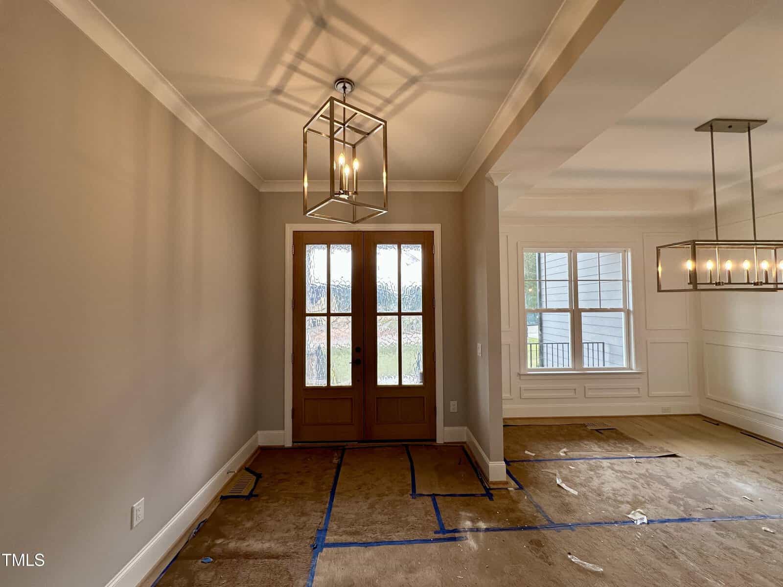 A room with unfinished floors, double wooden doors with frosted glass panels, beige walls, modern square light fixtures, and a large window with white trim. Blue tape marks are on the floor.