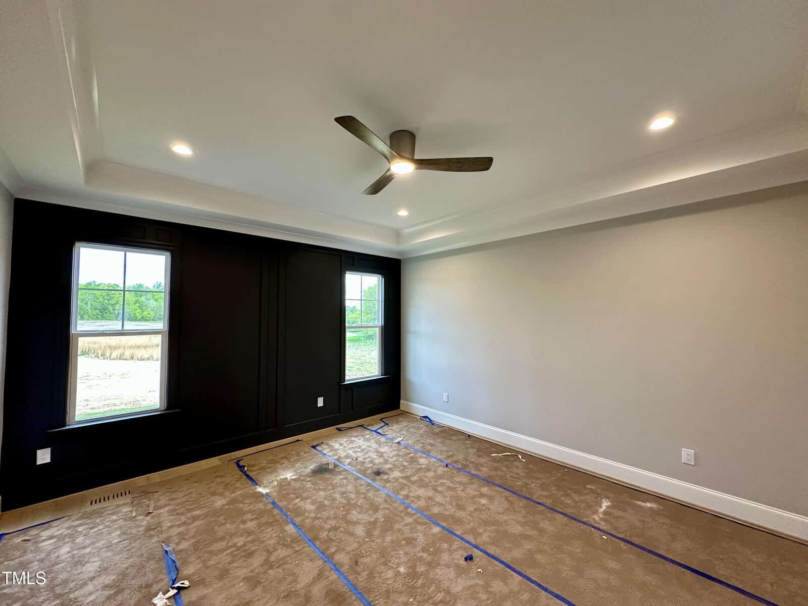 A modern, empty room with beige carpet, black accent wall, large windows, ceiling fan, and recessed lighting. Blue tape marks gridlines on the floor, and natural light comes through the windows.