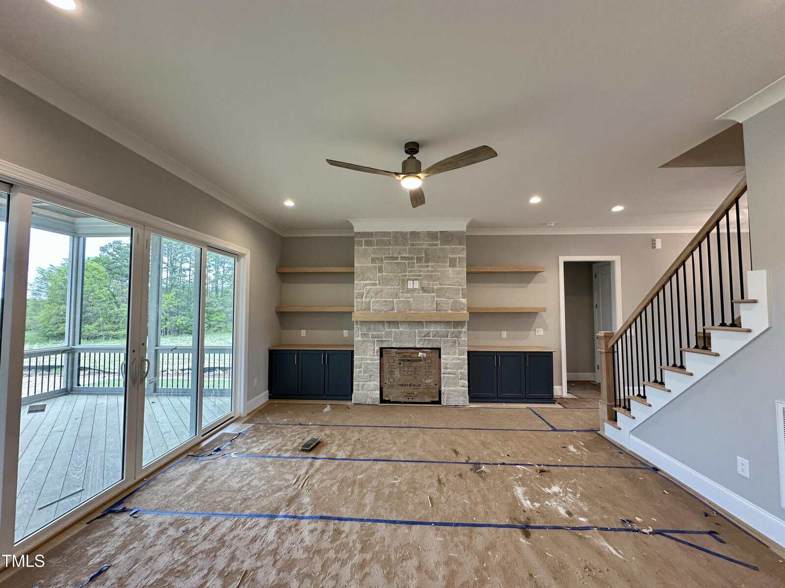 A bright living room features a stone fireplace, dark built-in cabinets, floating shelves, a ceiling fan, and large sliding doors leading to a deck. The floor is covered with protective paper. Stairs are visible to the right.