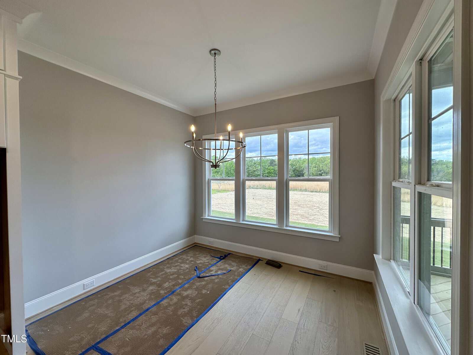 Bright dining room with large windows, modern chandelier, light wood floor, and neutral walls. Floor is partially covered for protection. Outdoor fields and trees visible through the windows.