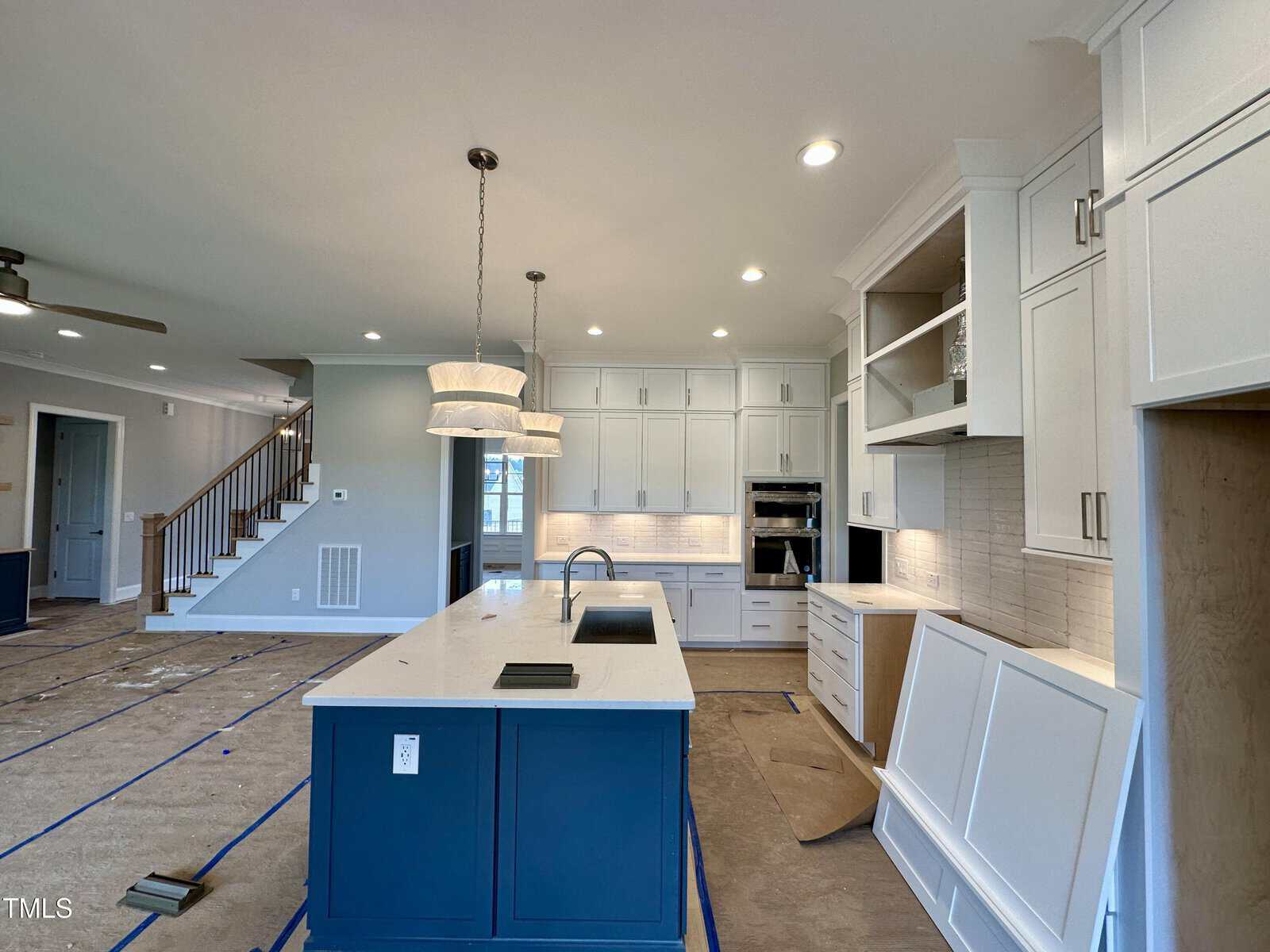 Modern kitchen with a large island featuring a sink and blue cabinets, white cabinetry throughout, pendant lights, and appliances built into the wall. Cardboard and blue tape cover parts of the floor, indicating ongoing construction.