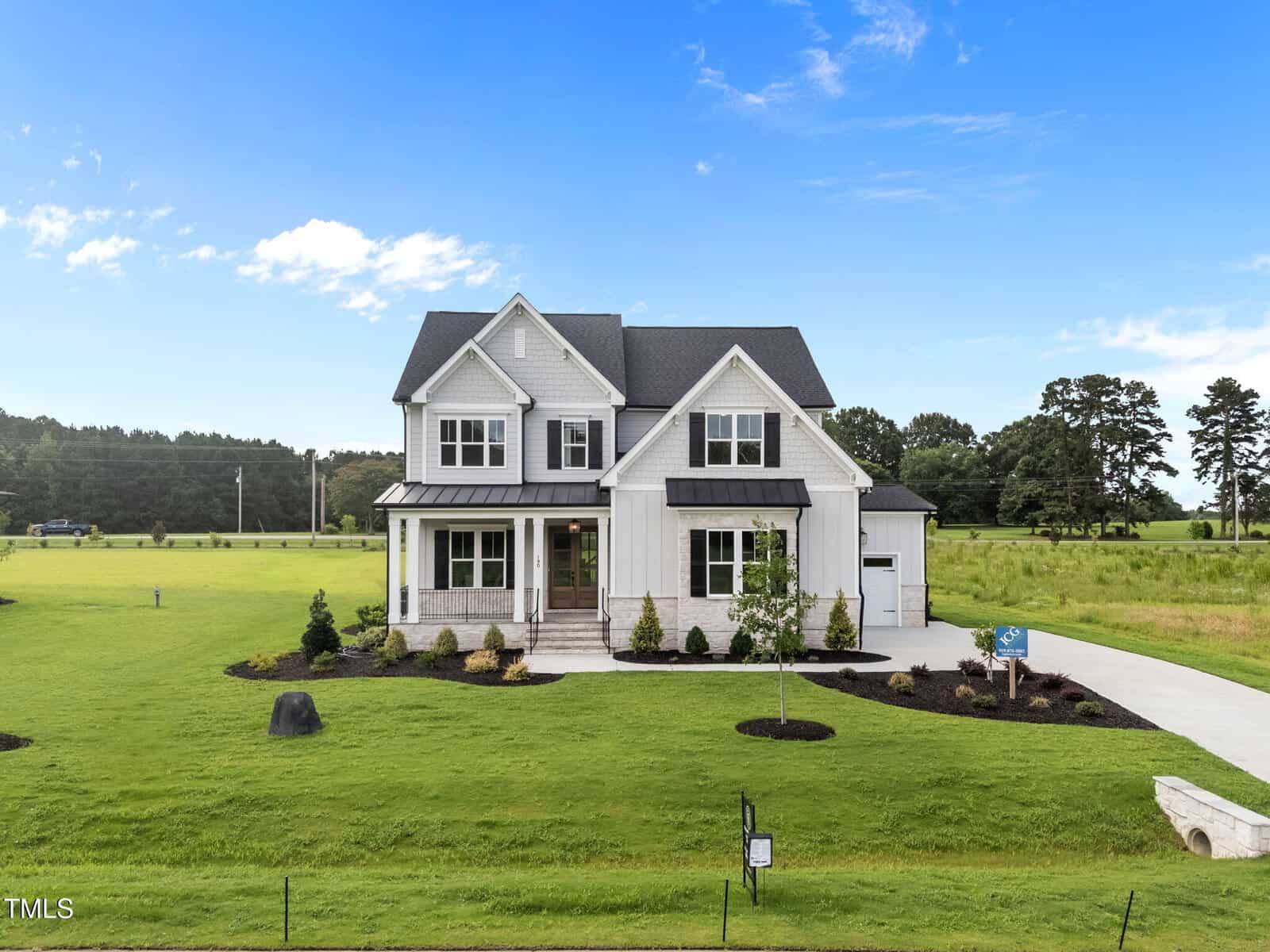Two-story modern farmhouse with light gray siding, black roof, and front porch, surrounded by a large, neatly manicured green lawn and landscaping, under a bright blue sky with a few clouds.